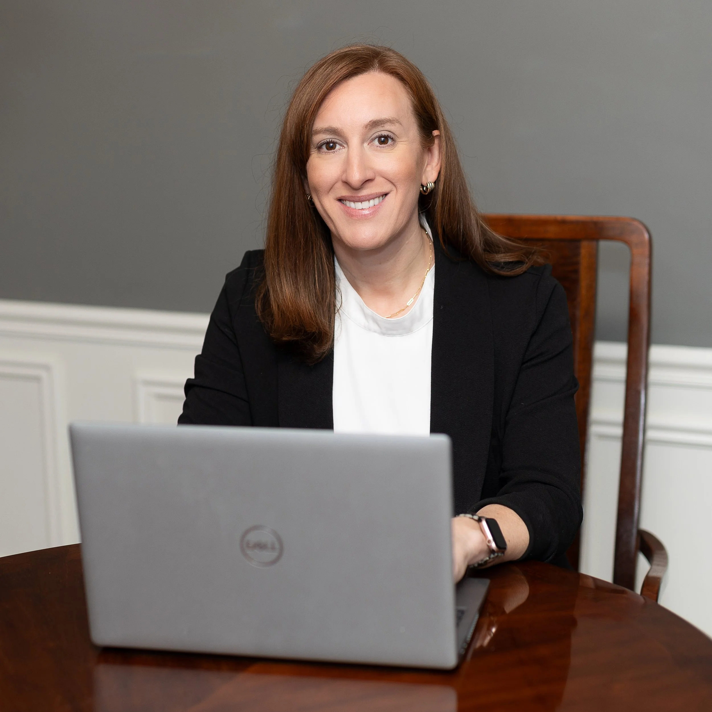 Dr. Tabatha Ritenour, wearing a black blazer and white shirt, is sitting at a table with a laptop in front of her. Dr. Ritenour is smiling, typing on a laptop, and looking at the camera, with a background that features a plain gray wall.