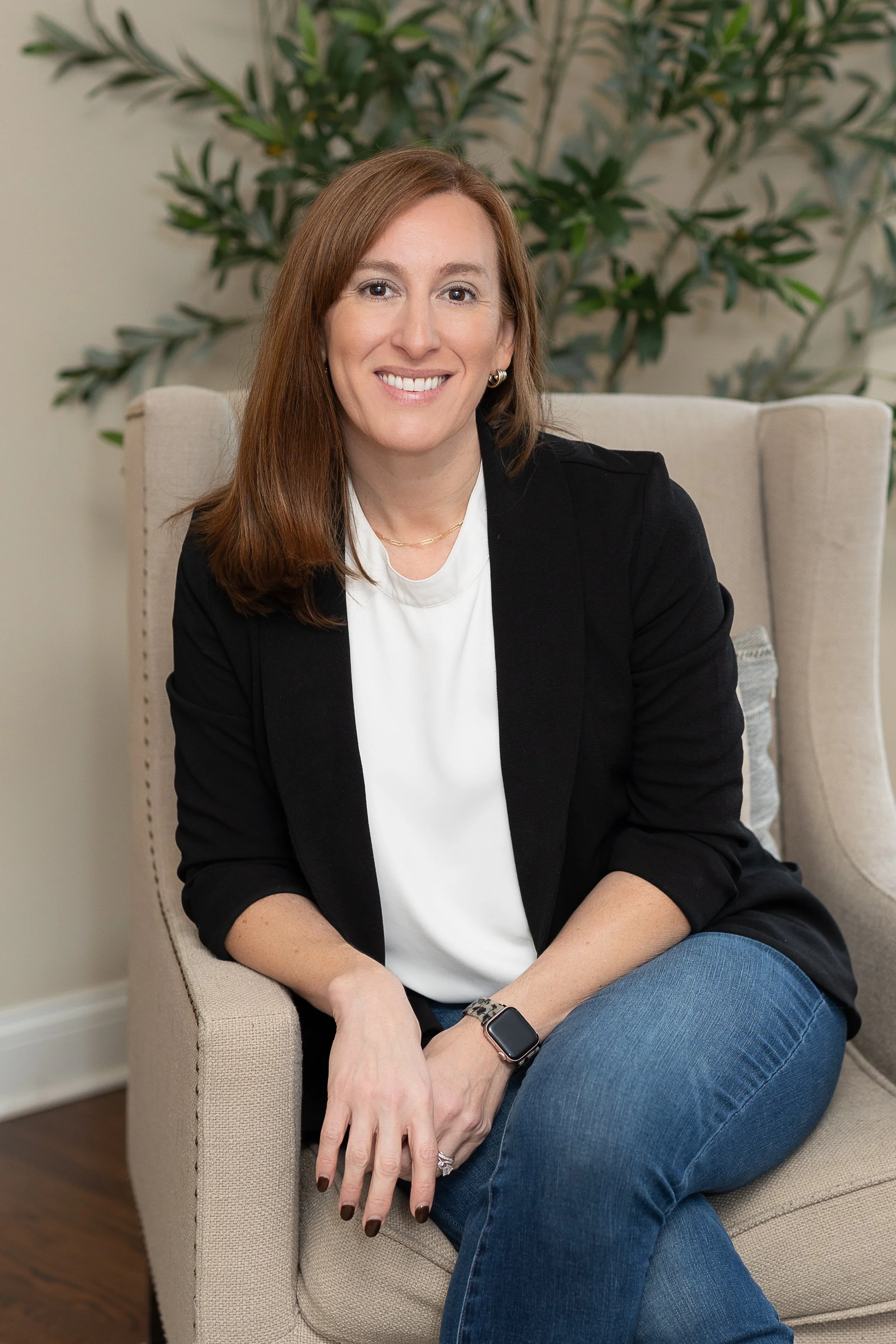 Dr. Tabatha Ritenour is sitting on a beige armchair in front of a leafy green plant, smiling at the camera. Dr. Ritenour is wearing a black blazer over a white top, blue jeans, and a smartwatch.