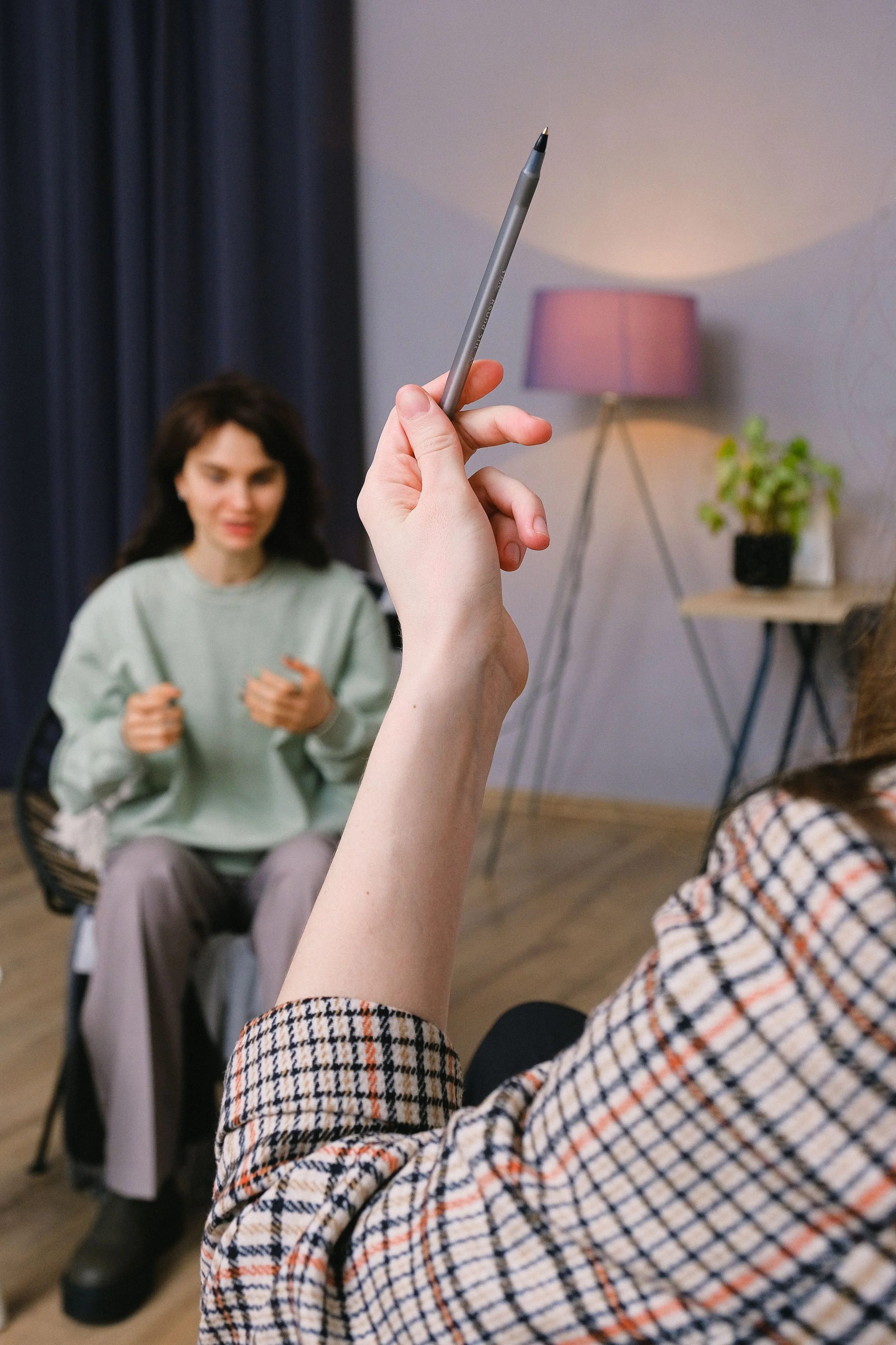 Person holding a pen in front of two women sitting in chairs indoors, with one woman in focus and the other slightly blurred, in a room with a lamp and a plant in the background.