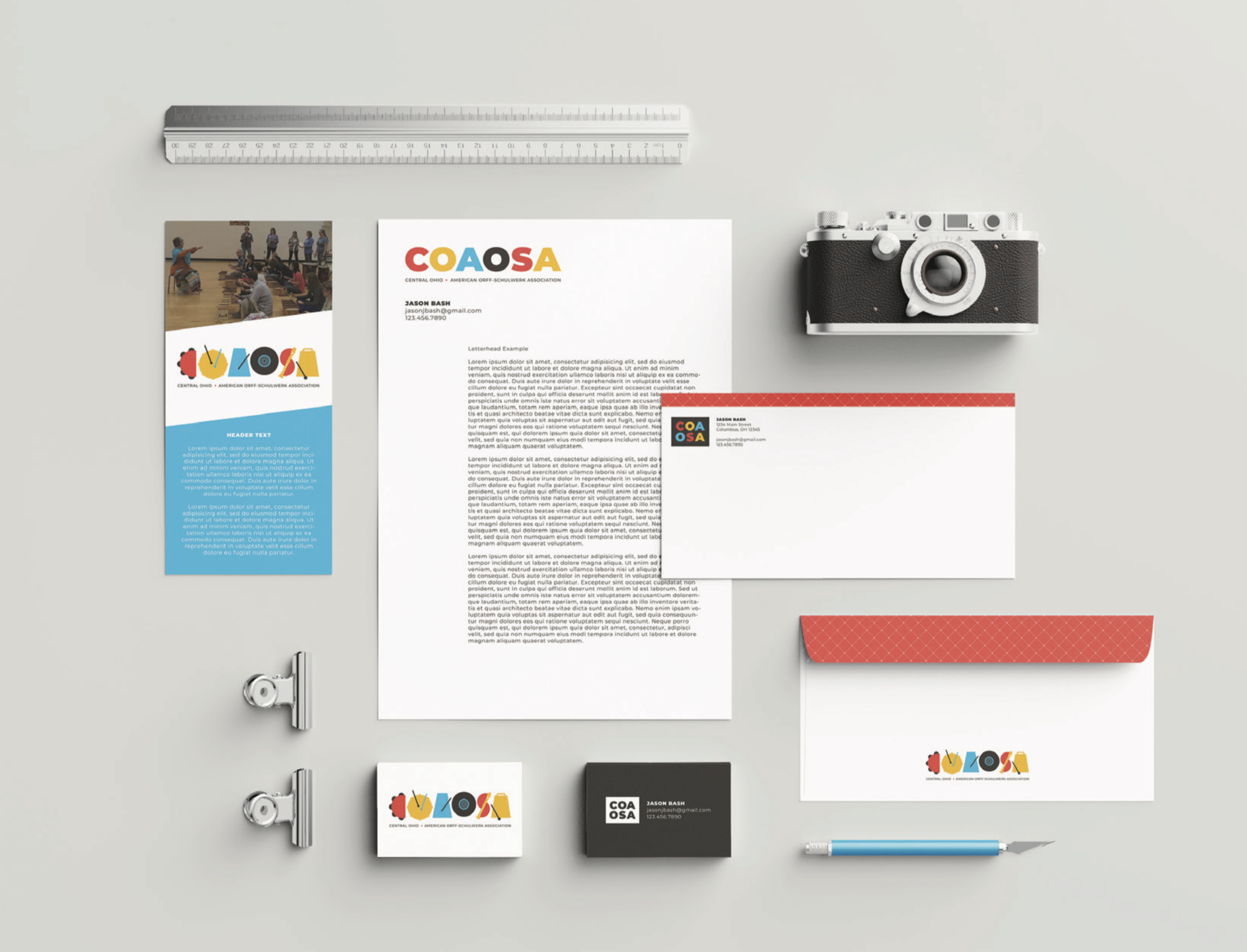 Flat lay of business branding items including a business card, a letter, a photograph, a notebook, binder clips, a pen, and a logo sticker, all arranged on a light gray background.