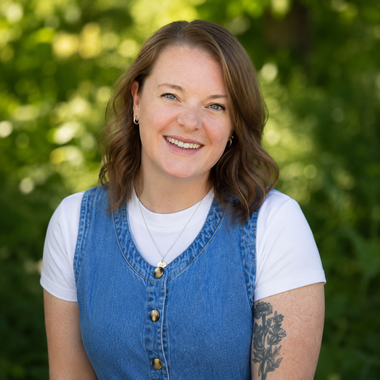 A woman with shoulder-length brown hair, wearing a white t-shirt and a blue denim vest, smiling outdoors with a blurred green leafy background.