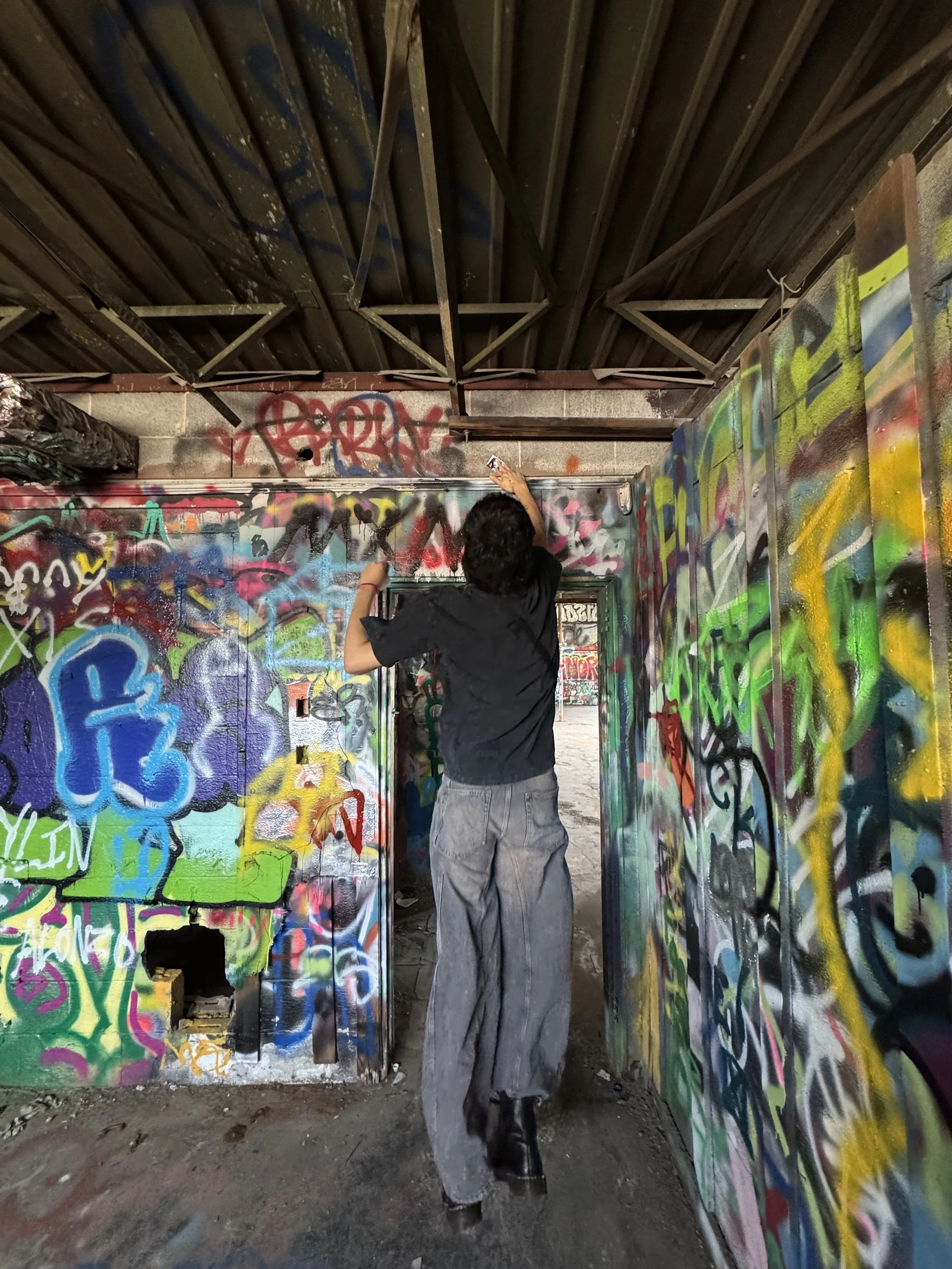 A person in black clothing is spray painting graffiti inside a structure with graffiti-covered walls and a metal roof.