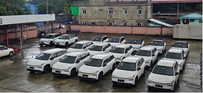 A parking lot with multiple white SUVs parked in rows on a wet concrete surface, surrounded by a pink wall and some buildings in the background.