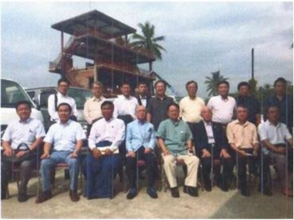 Group of men posing outdoors with a two-story wooden observation tower behind them and palm trees in the background.