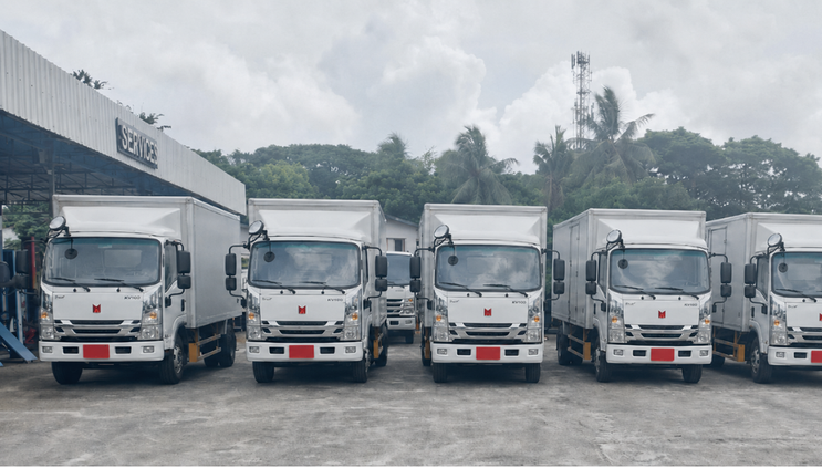 A row of white delivery trucks parked outside a service building with a sign, with trees and cloudy sky in the background.
