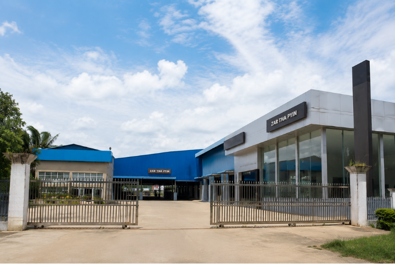 Commercial building with glass windows, a gated entrance, and signage that reads 'ZAR THA PYIN', surrounded by a concrete driveway and some green landscaping, under a cloudy sky.