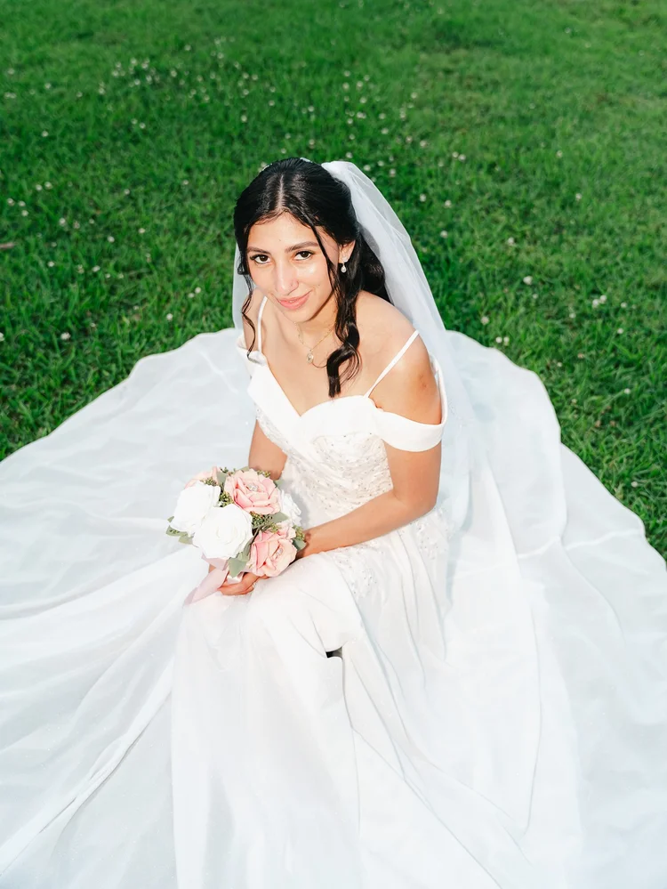 Bride in a white dress and veil sitting in the grass with her bouquet