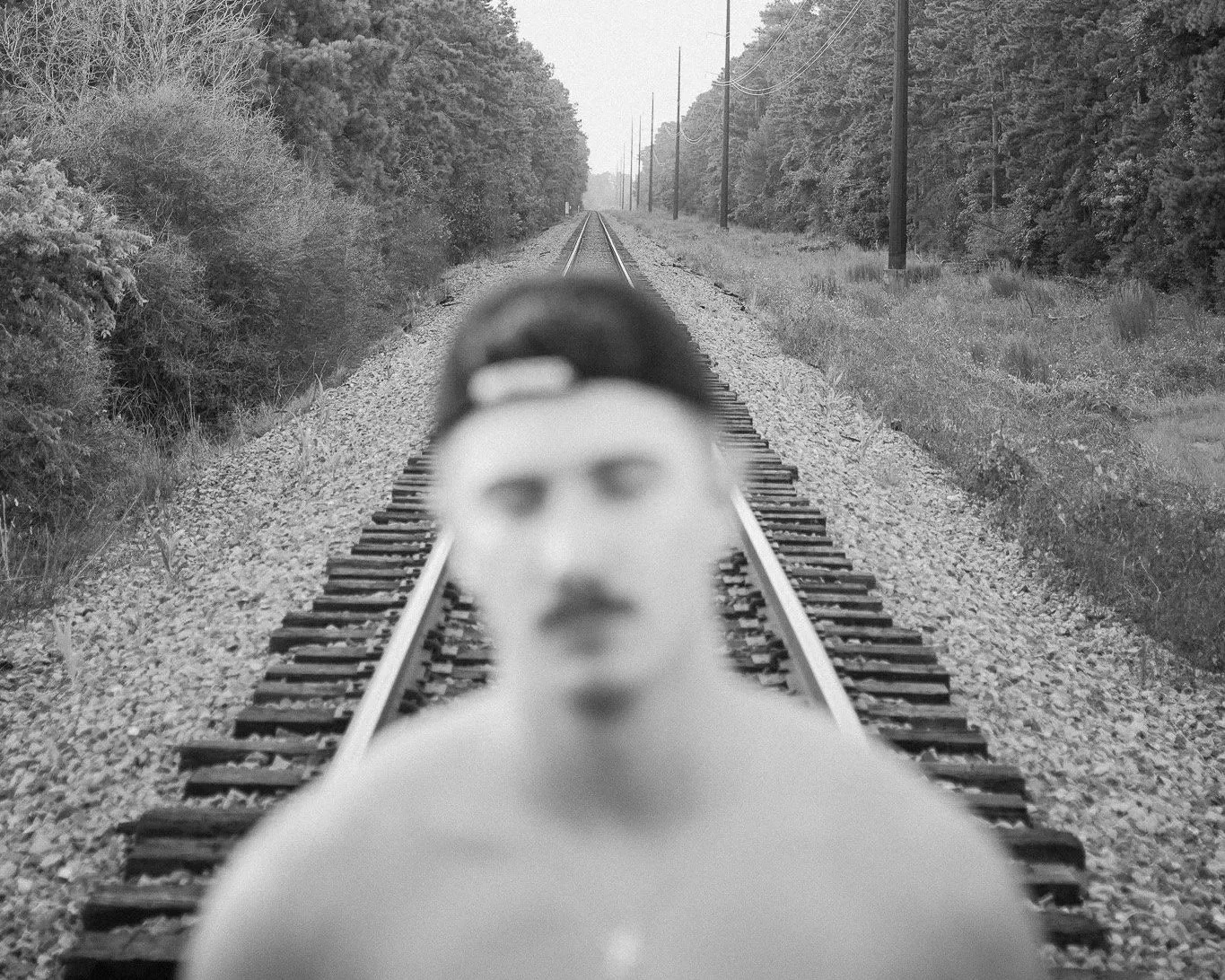 A black and white photo of a person with short hair, glasses, and lipstick standing on railway tracks. The person is out of focus, while the background of trees and power lines is clear.