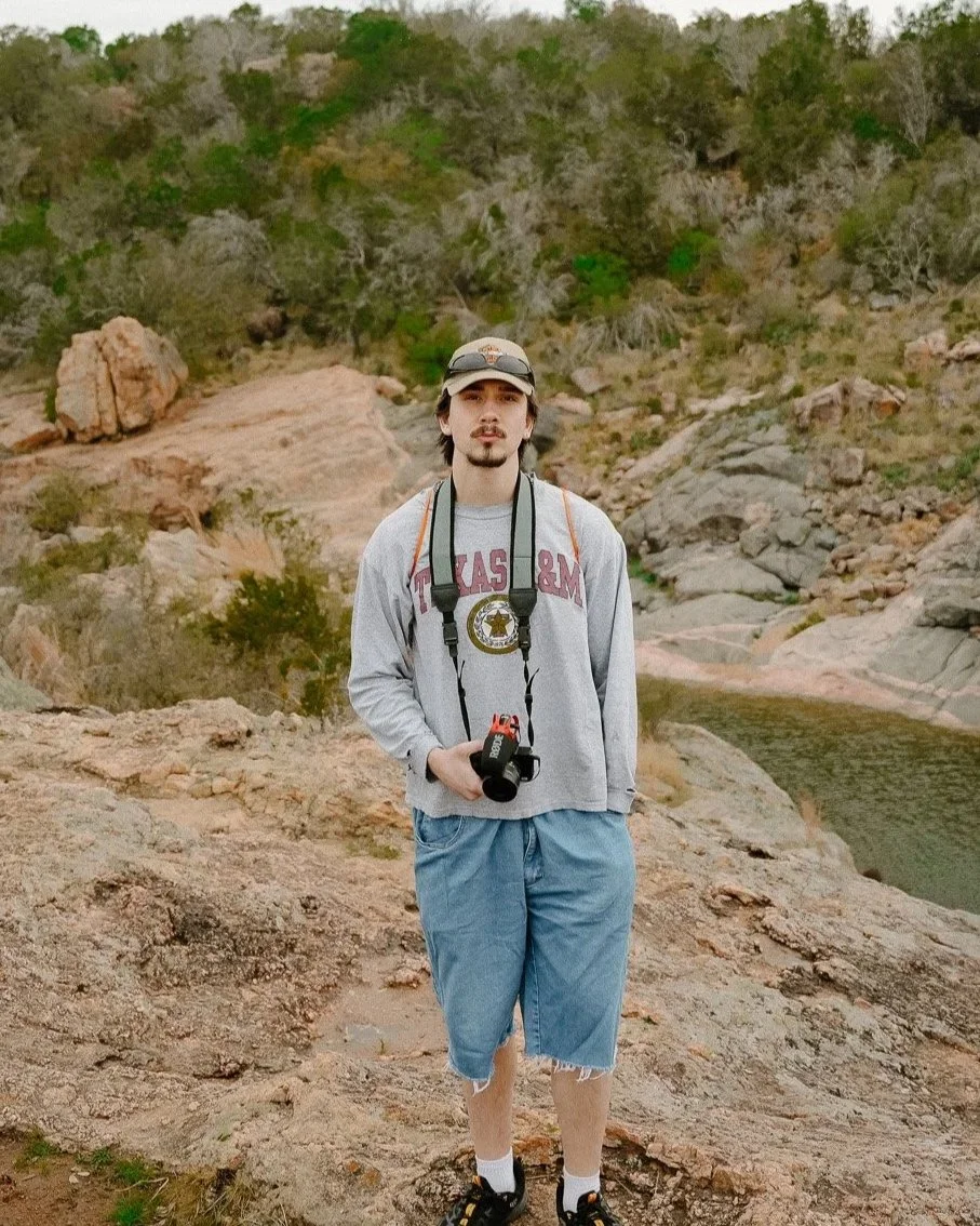 A young man with long brown hair, a mustache, and a goatee standing outdoors on rocky terrain, holding a camera with a strap around his neck, wearing a gray Texas A&M sweatshirt and denim shorts, in a natural landscape with trees and rocks in the background.