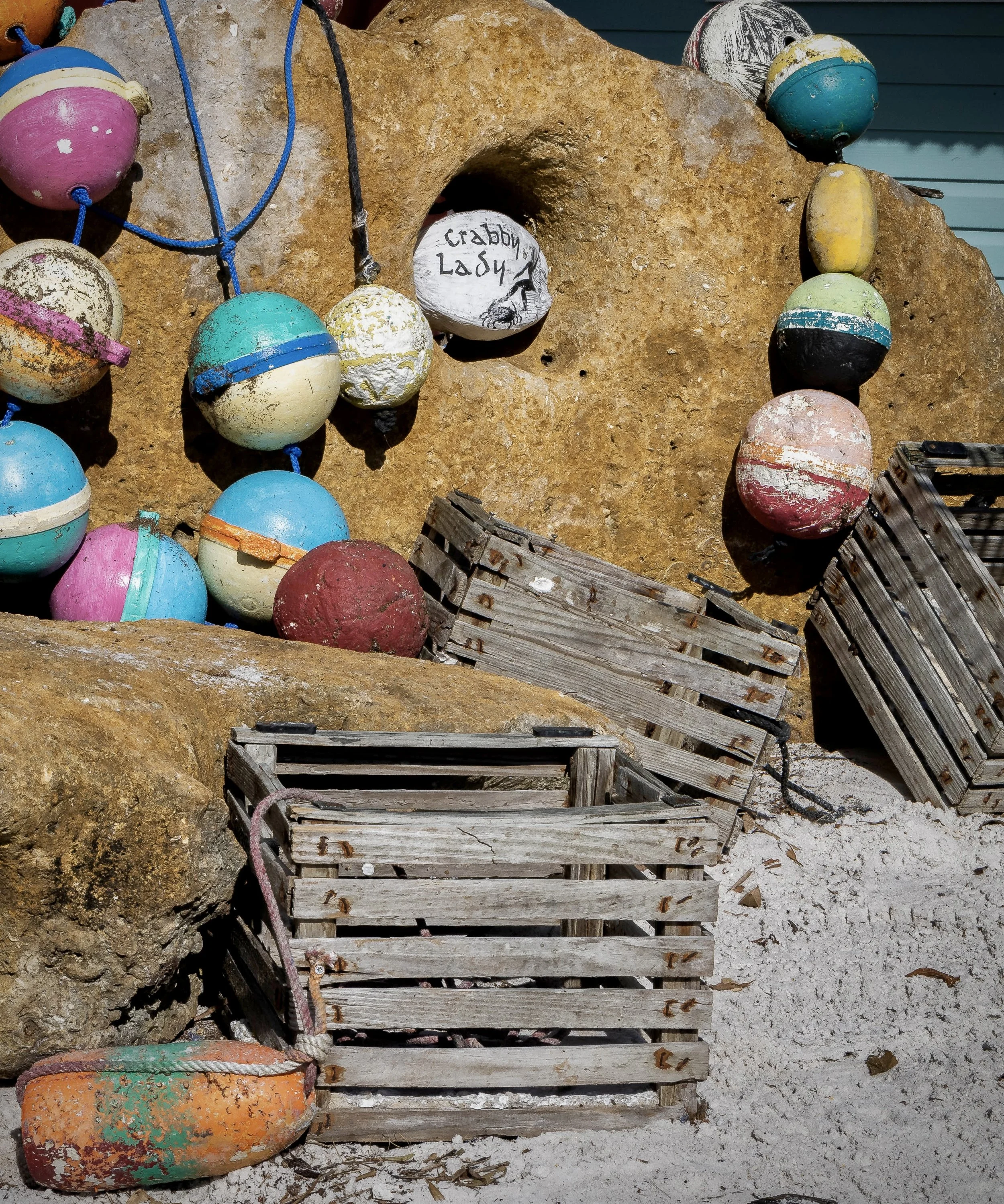 A collection of weathered, colorful buoys resting on a sandy surface, with some placed on or near a rustic wooden dock, against a rock background. There is a white rock inscribed with 'Crabby Lady' and a drawing of a crab.