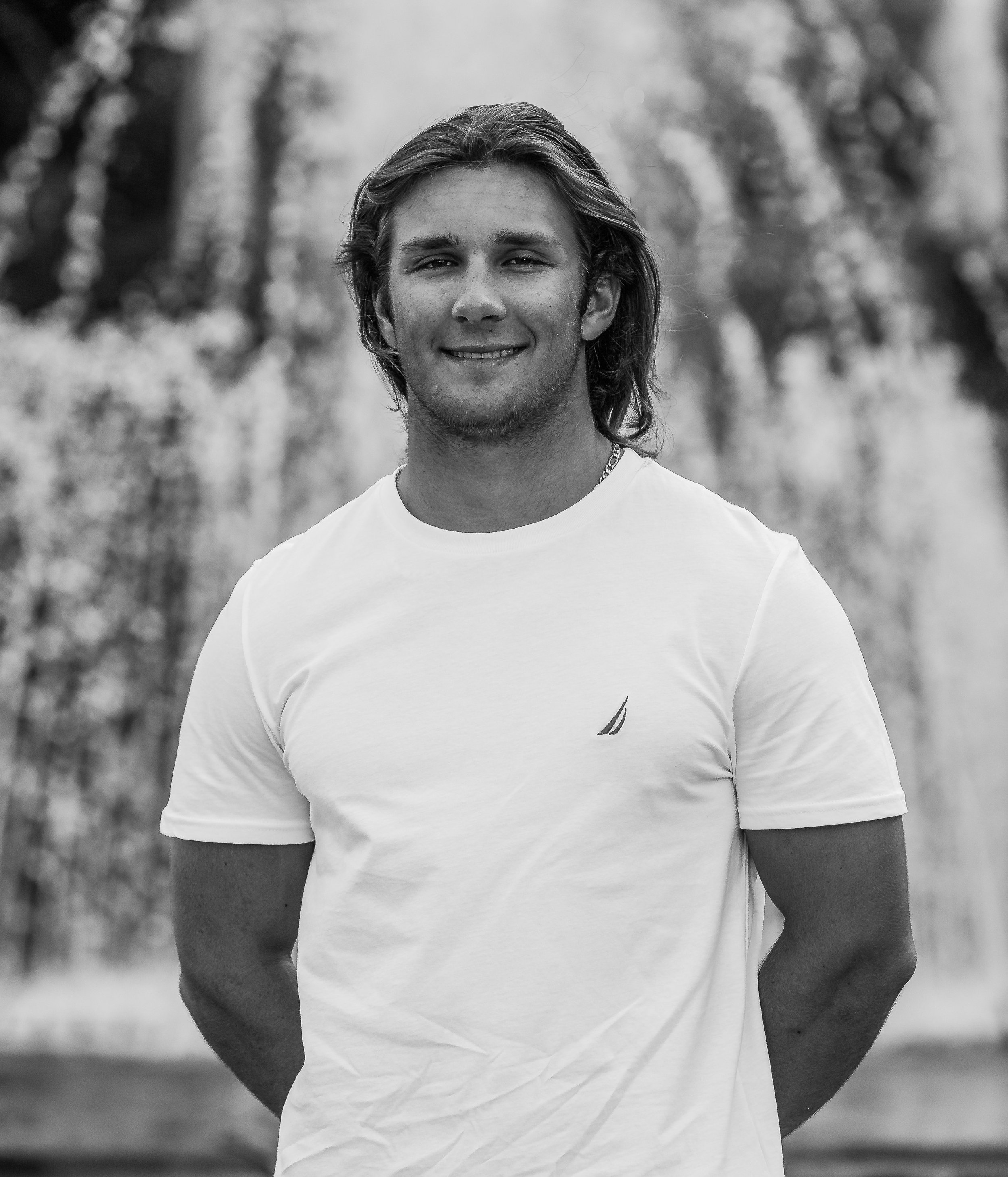 A young man with long hair, wearing a white t-shirt and a chain necklace, smiling outdoors.