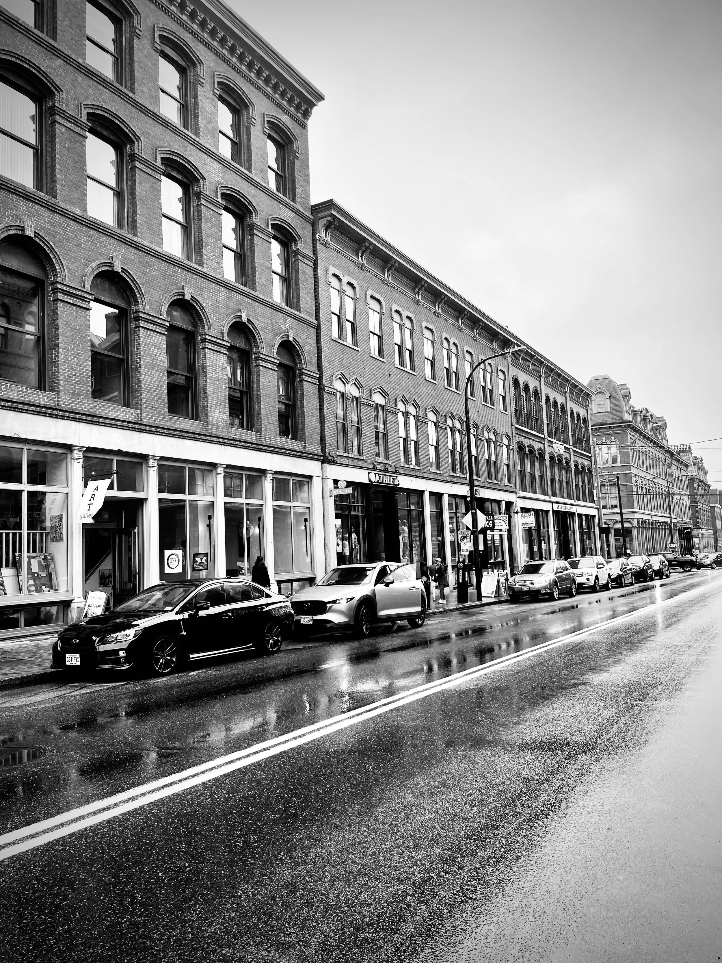 Black and white image of a city street with parked cars, wet pavement, and multi-story brick buildings with large windows.
