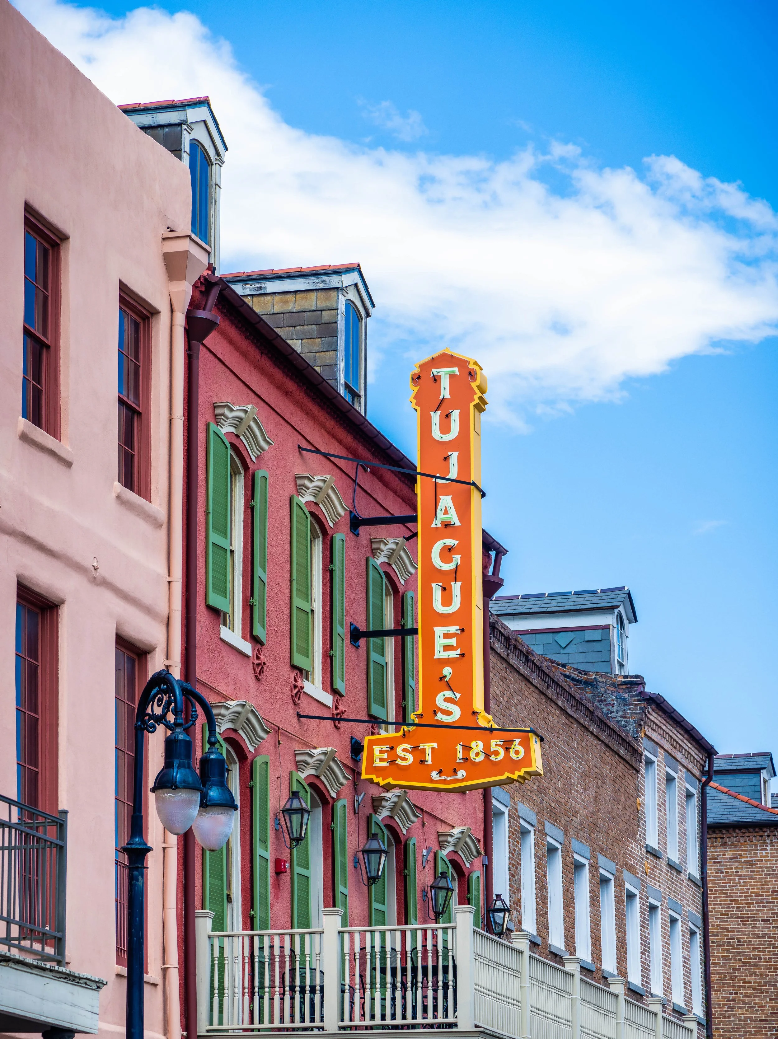 Colorful buildings with green shutters and a vertical sign reading "TUJACUES" with an additional sign below indicating "EST 1856" against a backdrop of a blue sky with some clouds.