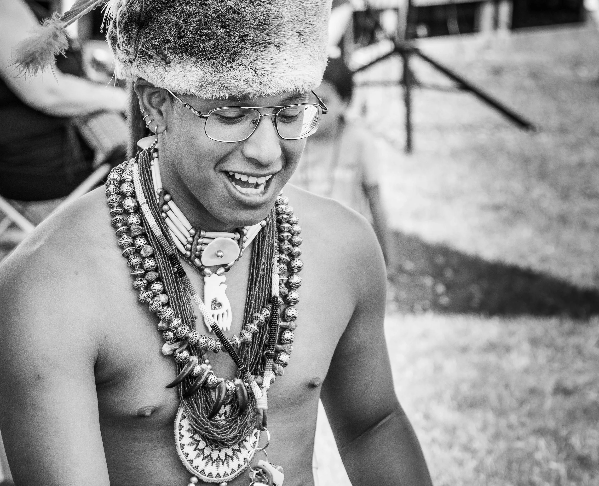 A person wearing multiple layered beaded necklaces, earrings, glasses, and a hat made of fur and animal skin. The person appears to be smiling and looking down, with an outdoor background.