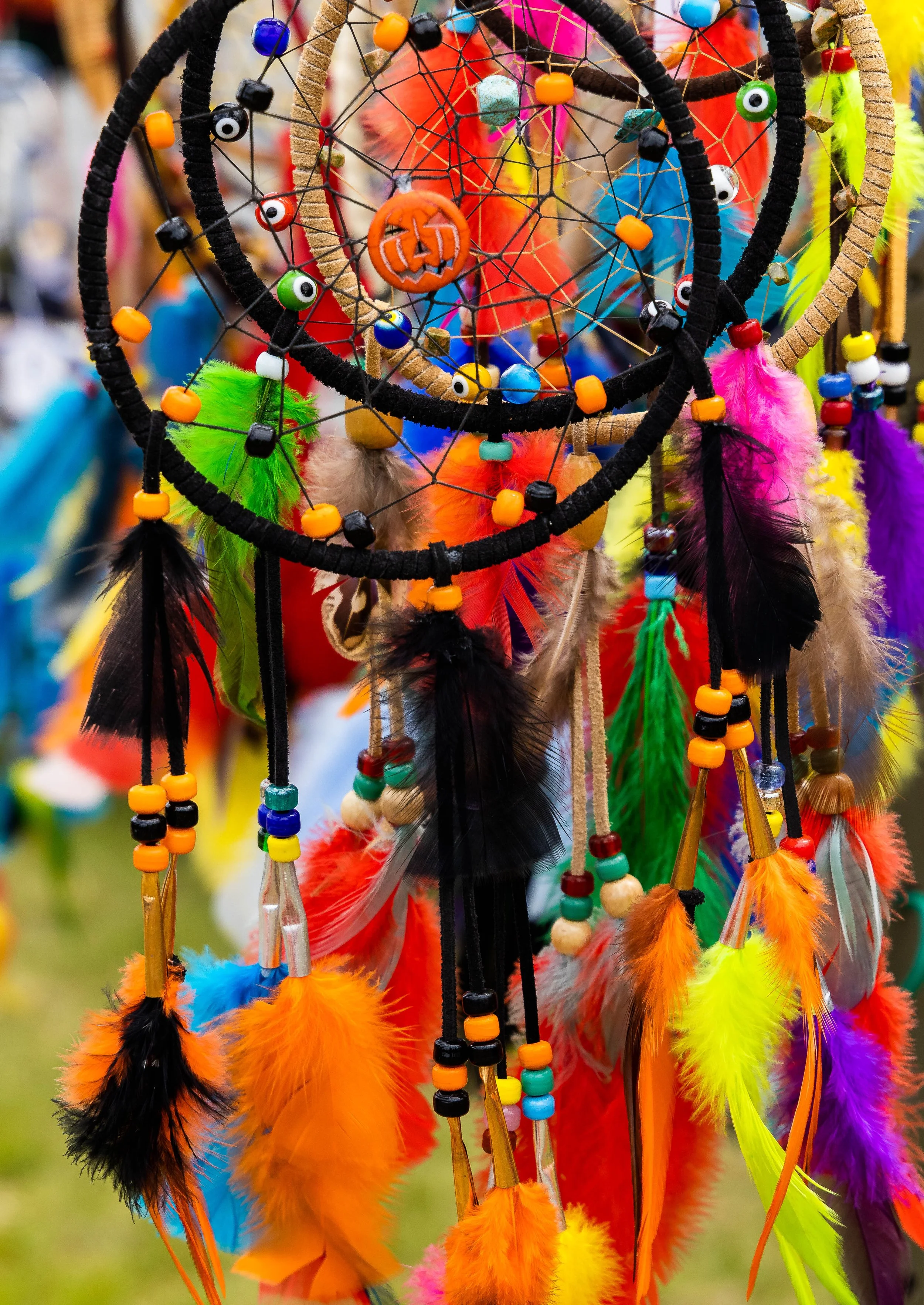 Colorful dreamcatchers with feathers and beads hanging in a display, featuring vibrant orange, green, yellow, pink, purple, and black feathers, with some feathers decorated with beads and netting.