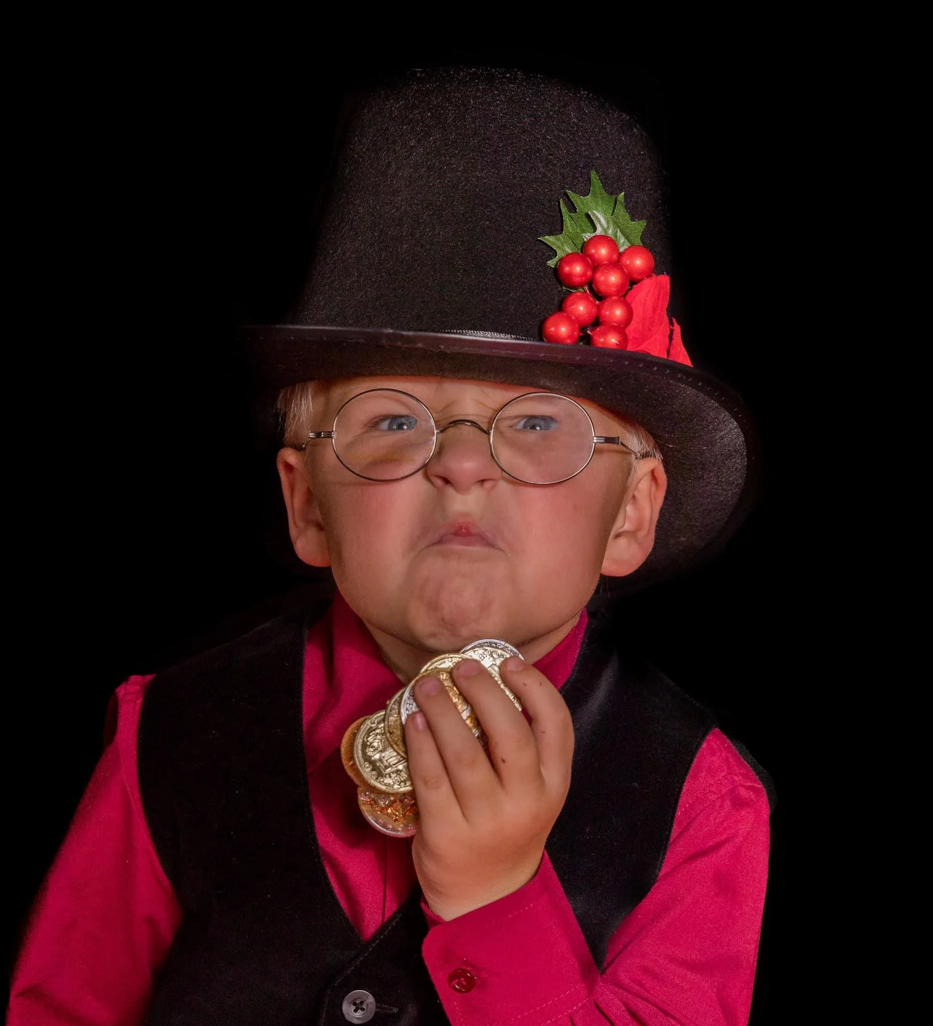 A person wearing a black top hat decorated with red berries, green leaves, and a red ribbon, along with a pink shirt and black vest, holding several gold coins with their face partially visible.