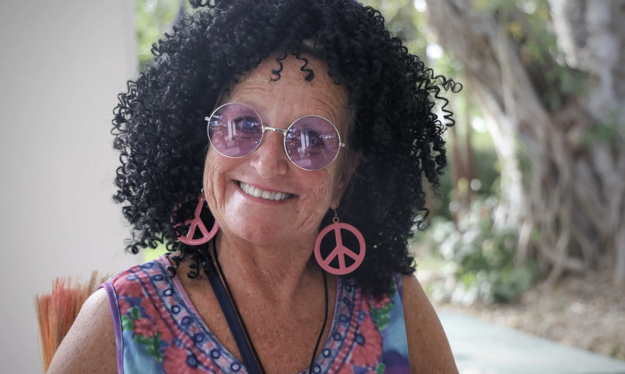 A smiling woman with curly black hair wearing pink peace sign earrings, purple round sunglasses, and a colorful sleeveless top. The background is outdoors with trees and greenery.