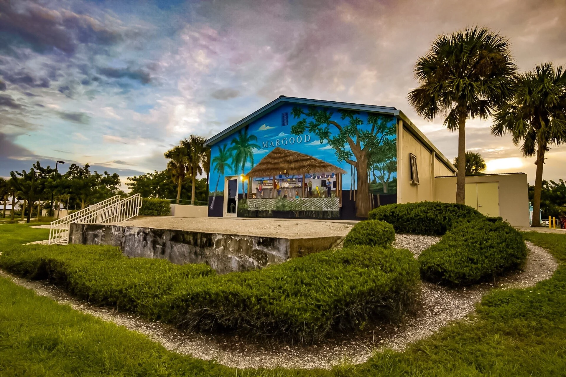 A building with a mural of a tropical scene including palm trees and a thatched hut, with the word 'MARGOAD' on it, surrounded by green plants and trees, during sunset.