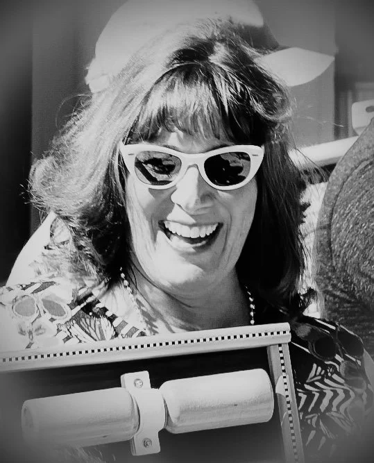 A woman with dark hair wearing white sunglasses and a hat, smiling outdoors.