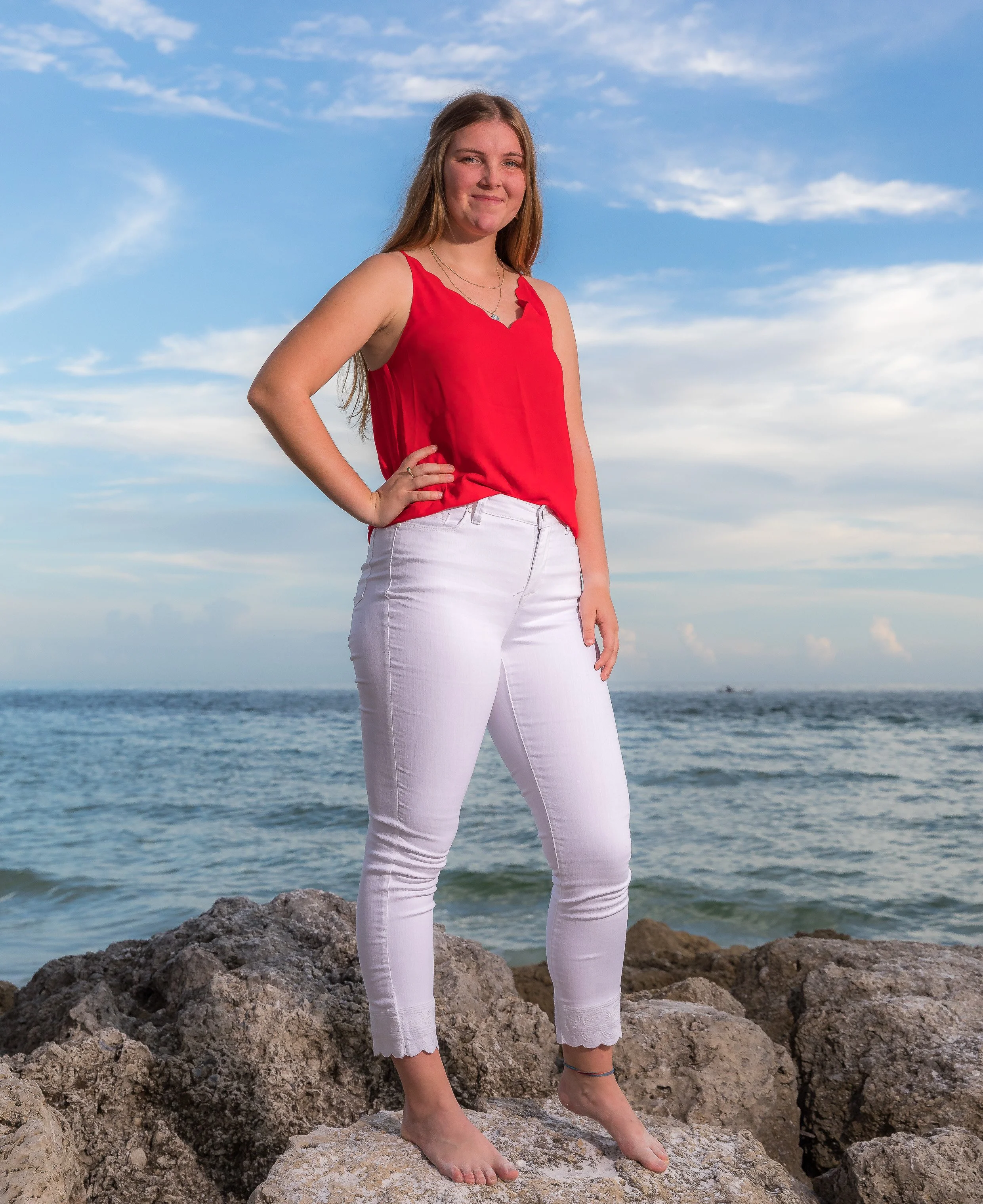 Young woman standing barefoot on rocks by the ocean, wearing a red sleeveless top and white pants, with her hand on her hip, smiling at the camera under a blue sky with clouds.