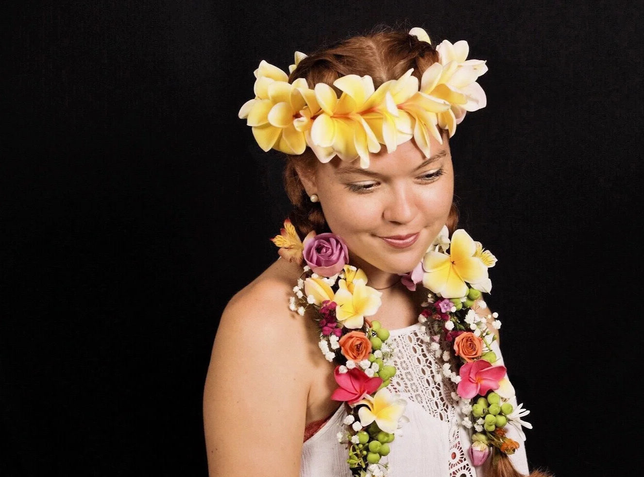 A woman wearing a floral lei and headpiece with yellow, pink, purple, and white flowers, against a black background.