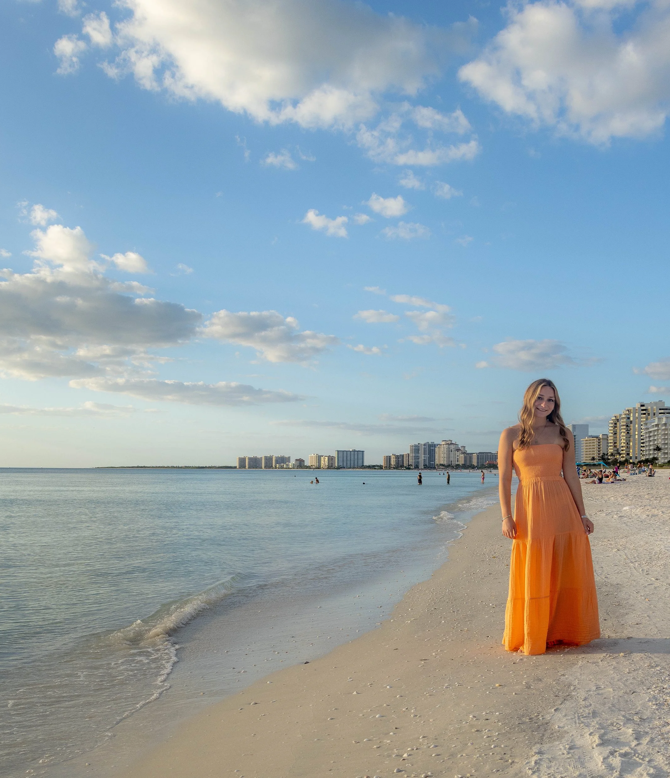 A young woman in a strapless orange dress standing on a beach with a city skyline in the background, under a sky with scattered clouds.