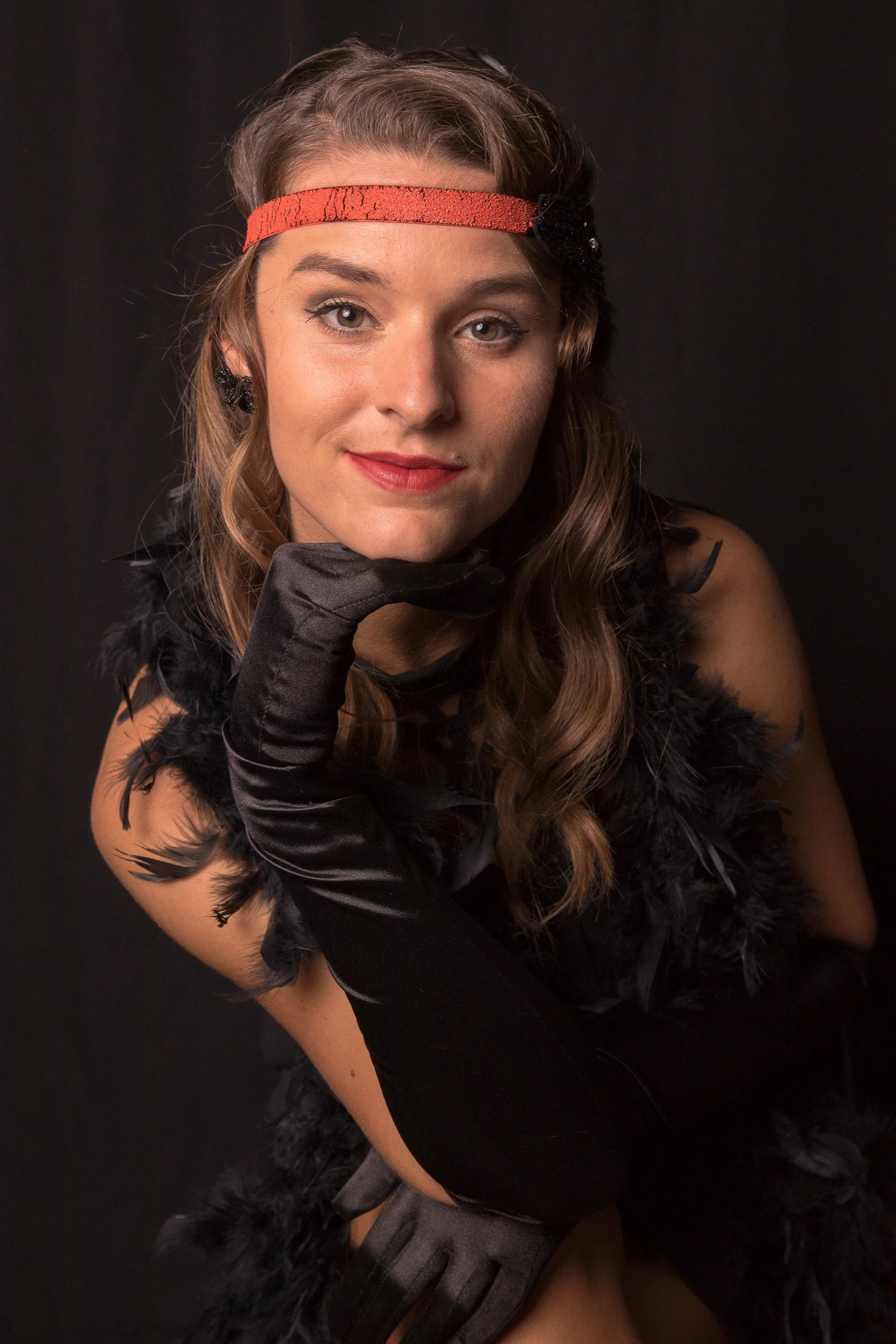 A woman with long wavy brown hair and light skin, wearing a red headband, black feathered costume, black gloves, and earrings, posing against a black background.