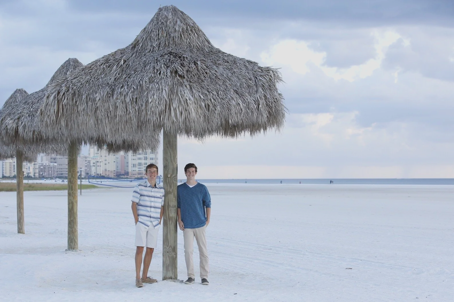 Two young men standing under a large thatched beach umbrella on a white sandy beach, with a city skyline in the background and a cloudy sky.