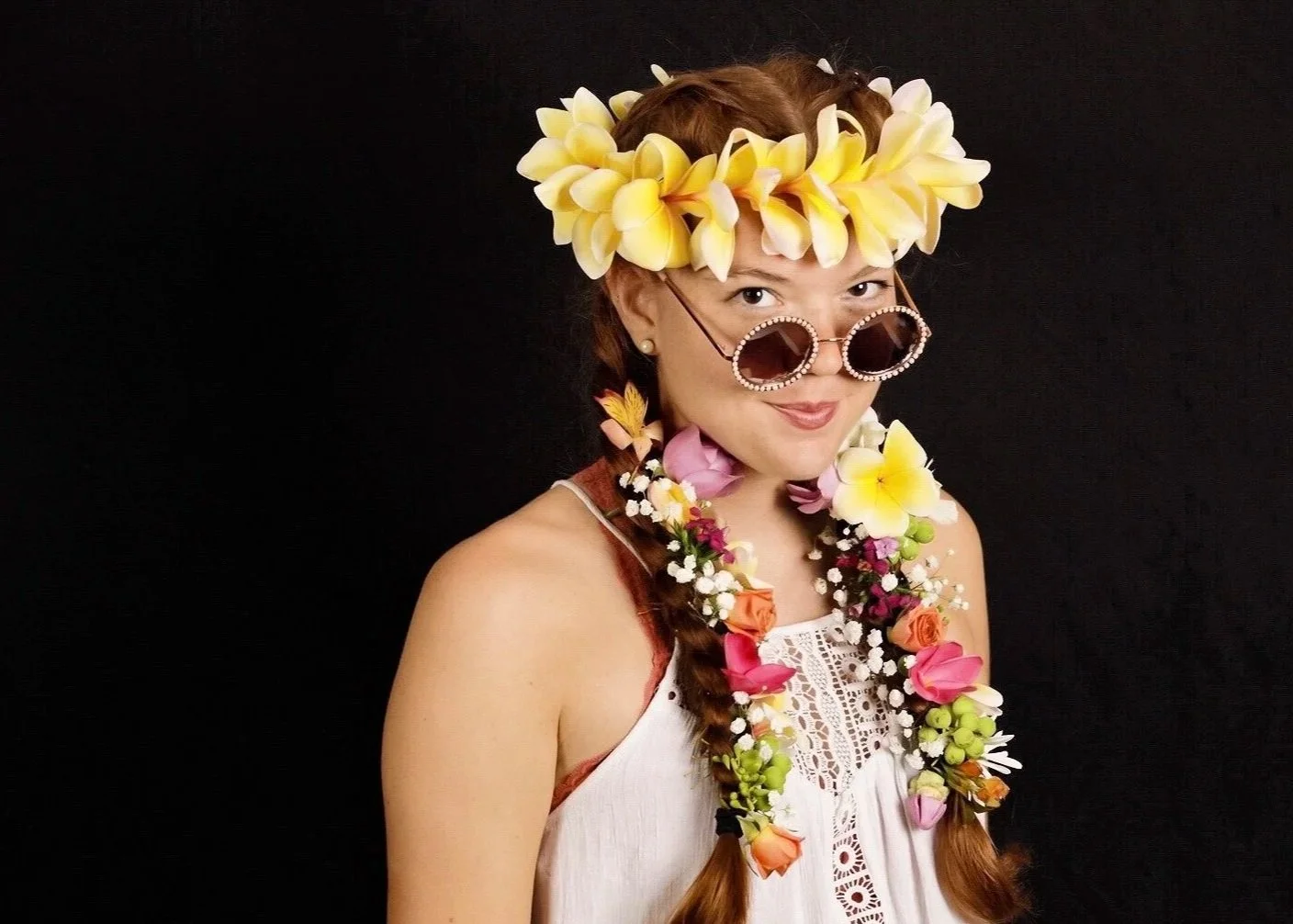 A woman wearing a flower crown, sunglasses, and a floral lei against a black background.