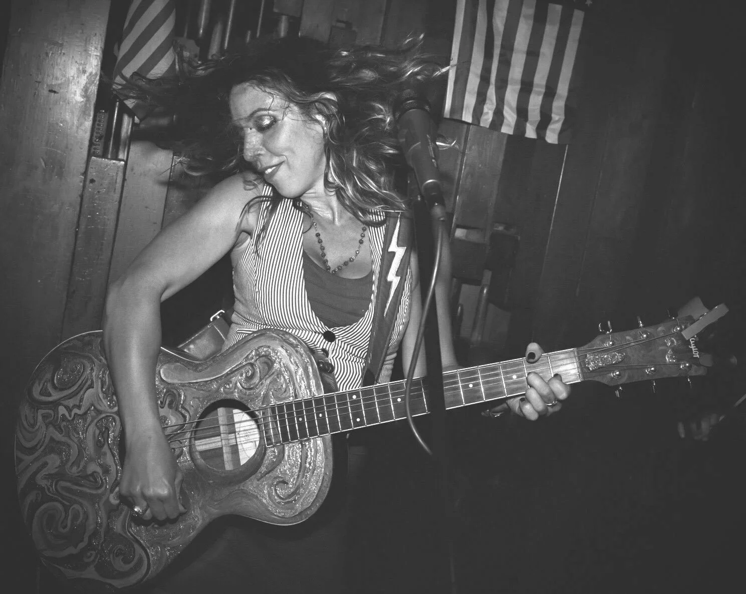 A woman singing and playing an acoustic guitar with a floral design in a dimly lit, rustic setting, possibly a bar or club, with American flags in the background.