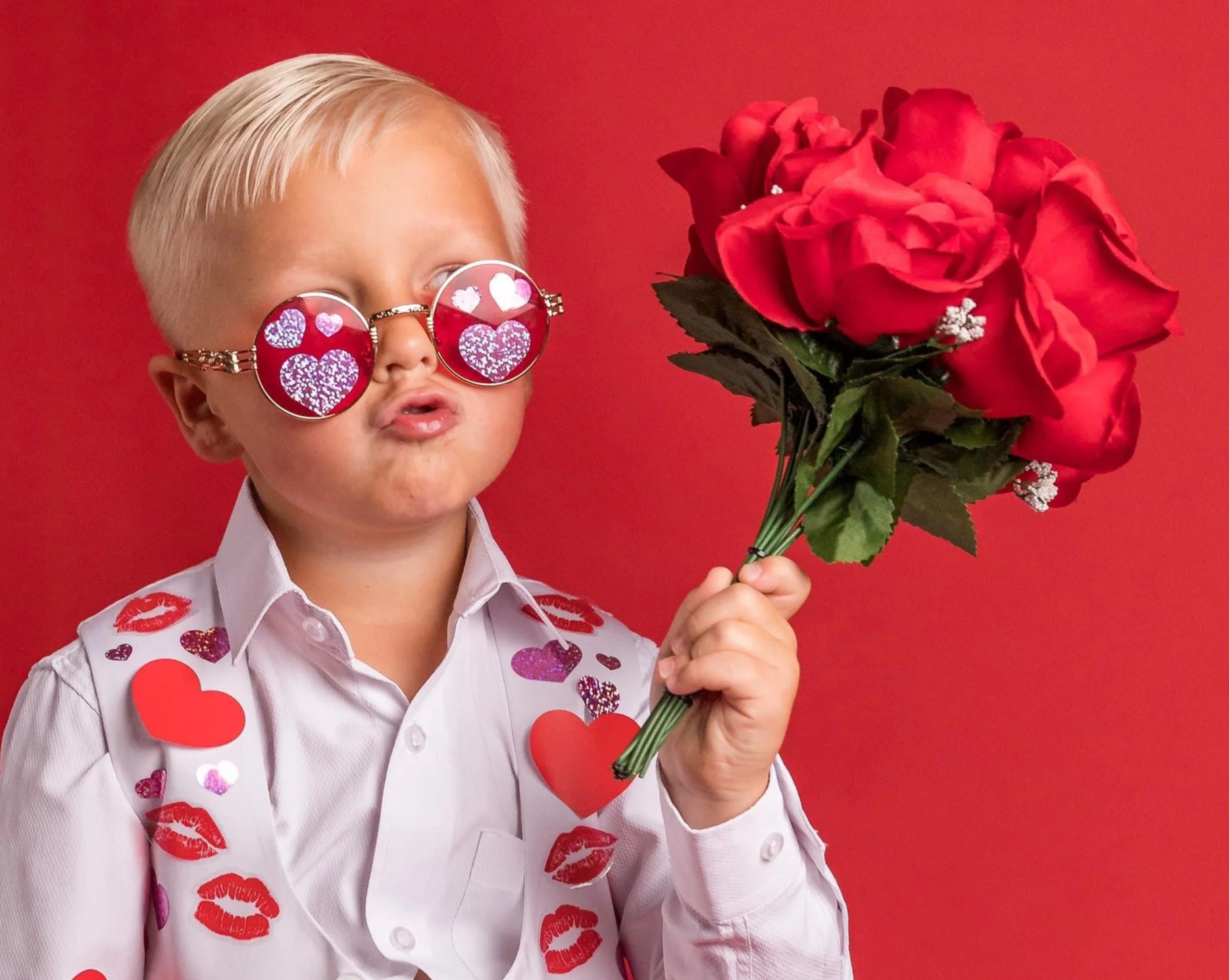 A young boy with blonde hair wearing sunglasses with heart-shaped reflections. He is holding a bouquet of red roses and is dressed in a white shirt decorated with red and purple heart patterns. The background is solid red.
