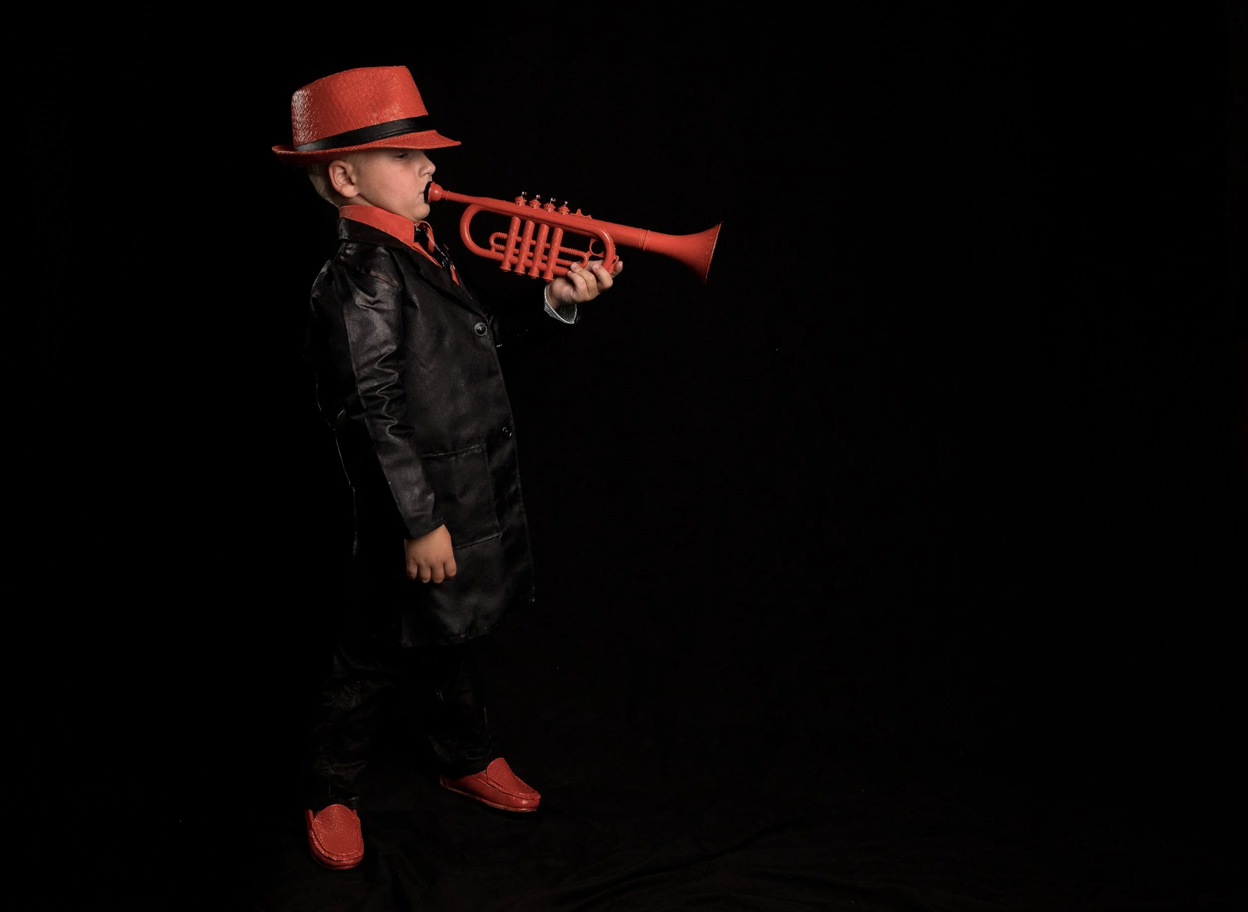 A young boy wearing a red hat, red shoes, and a black jacket, is holding a red toy trumpet to his mouth against a black background.