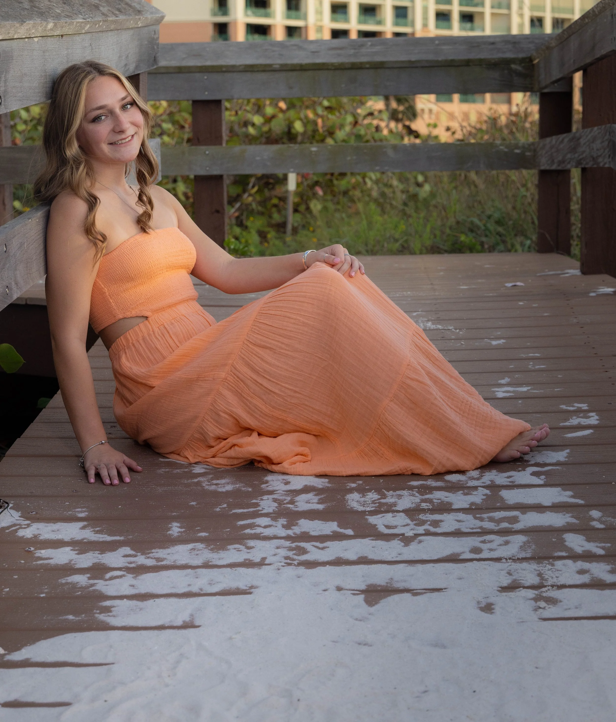 A young woman with wavy, shoulder-length blond hair sitting on a wooden deck, smiling and looking at the camera. She wears an orange strapless dress and a silver bracelet. The background includes a wooden railing and a building with balconies.