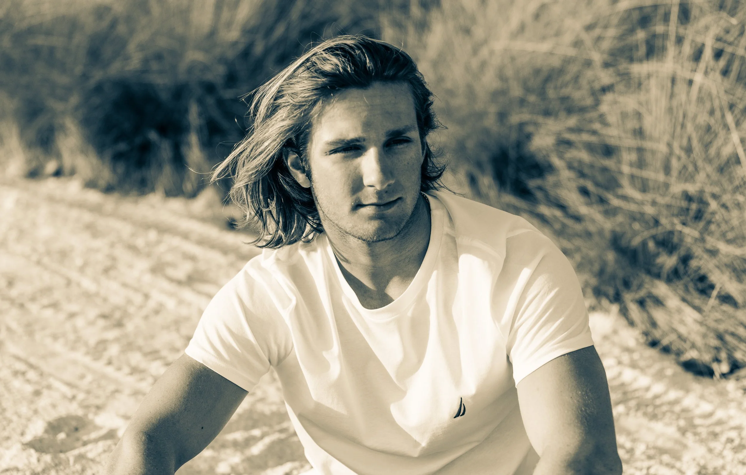 A young man with shoulder-length hair gazing into the distance outdoors on a sandy or rocky terrain with wild grass in the background, wearing a white t-shirt.