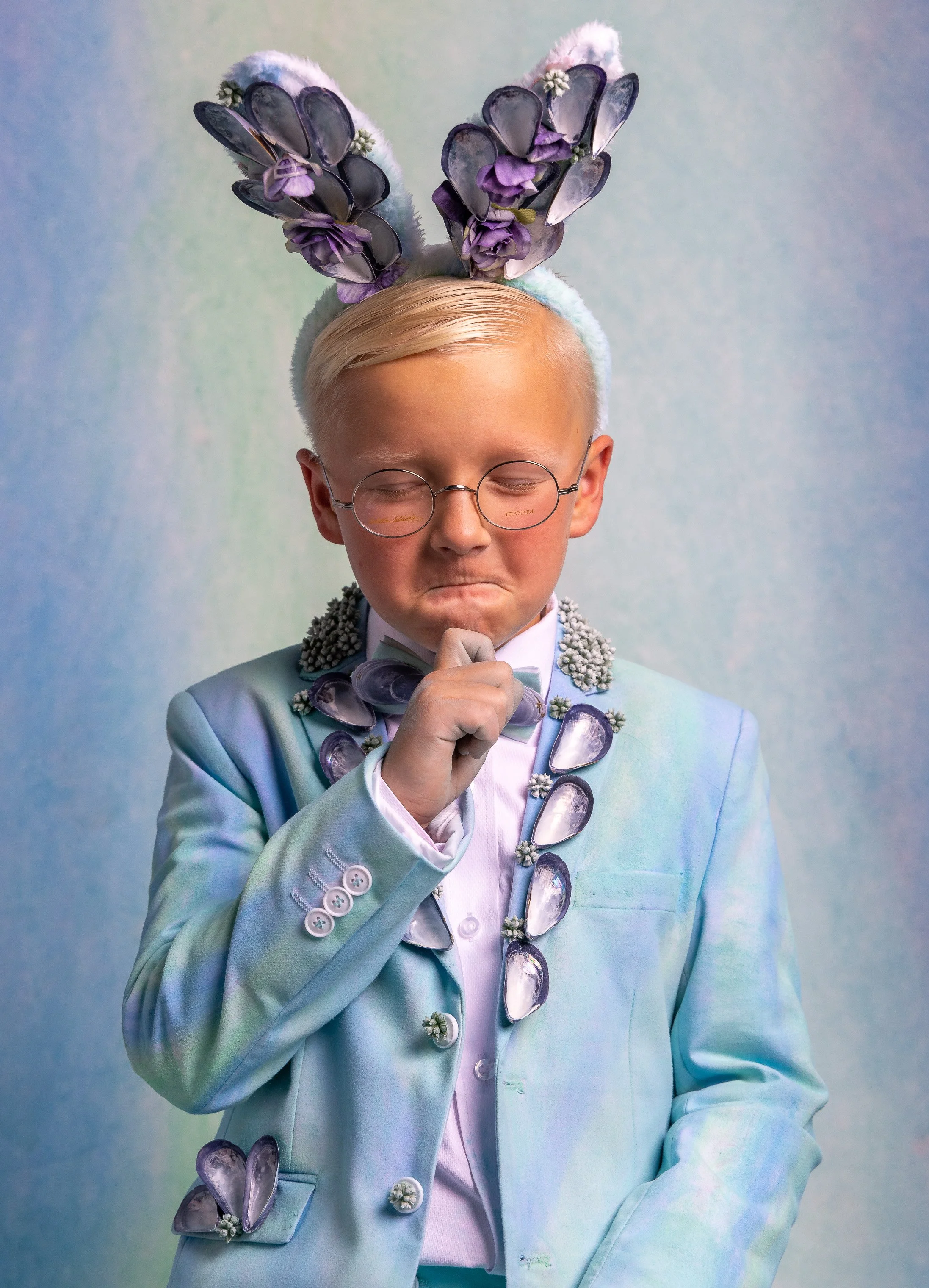 A person with platinum blonde hair, round glasses, and a thoughtful expression, wearing a pastel blue tuxedo with shell-shaped accessories and a bunny ear headband decorated with flowers and shells.