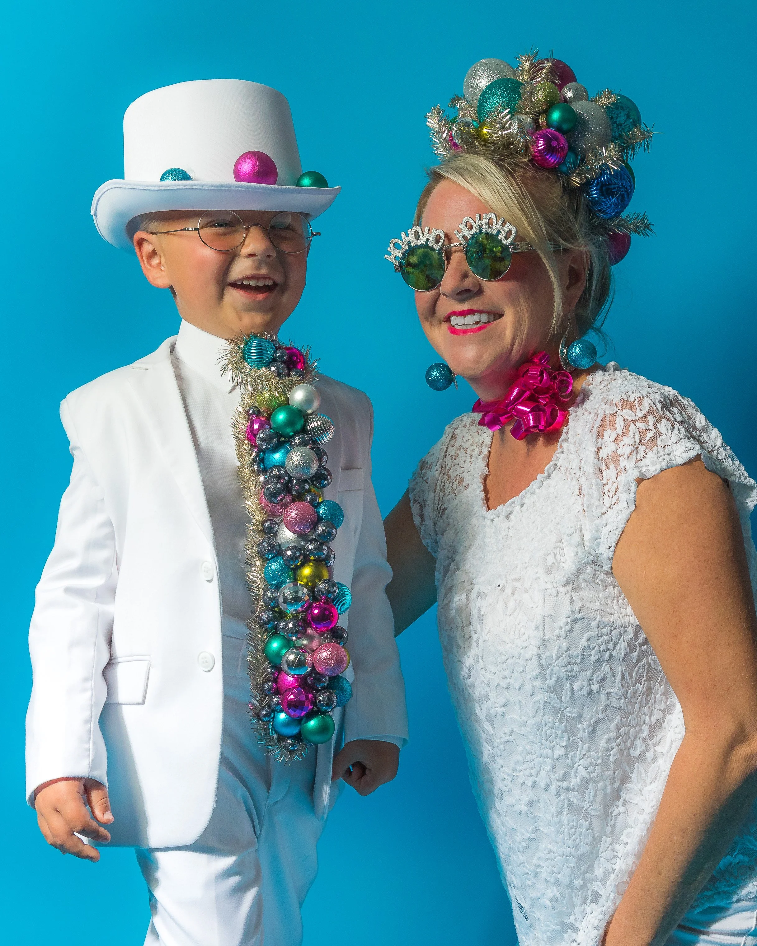 A woman and a boy dressed in holiday attire with festive accessories, standing against a bright blue background. The boy wears a white suit with a large white top hat and glasses, decorated with colorful ornaments and tinsel. The woman wears a white 