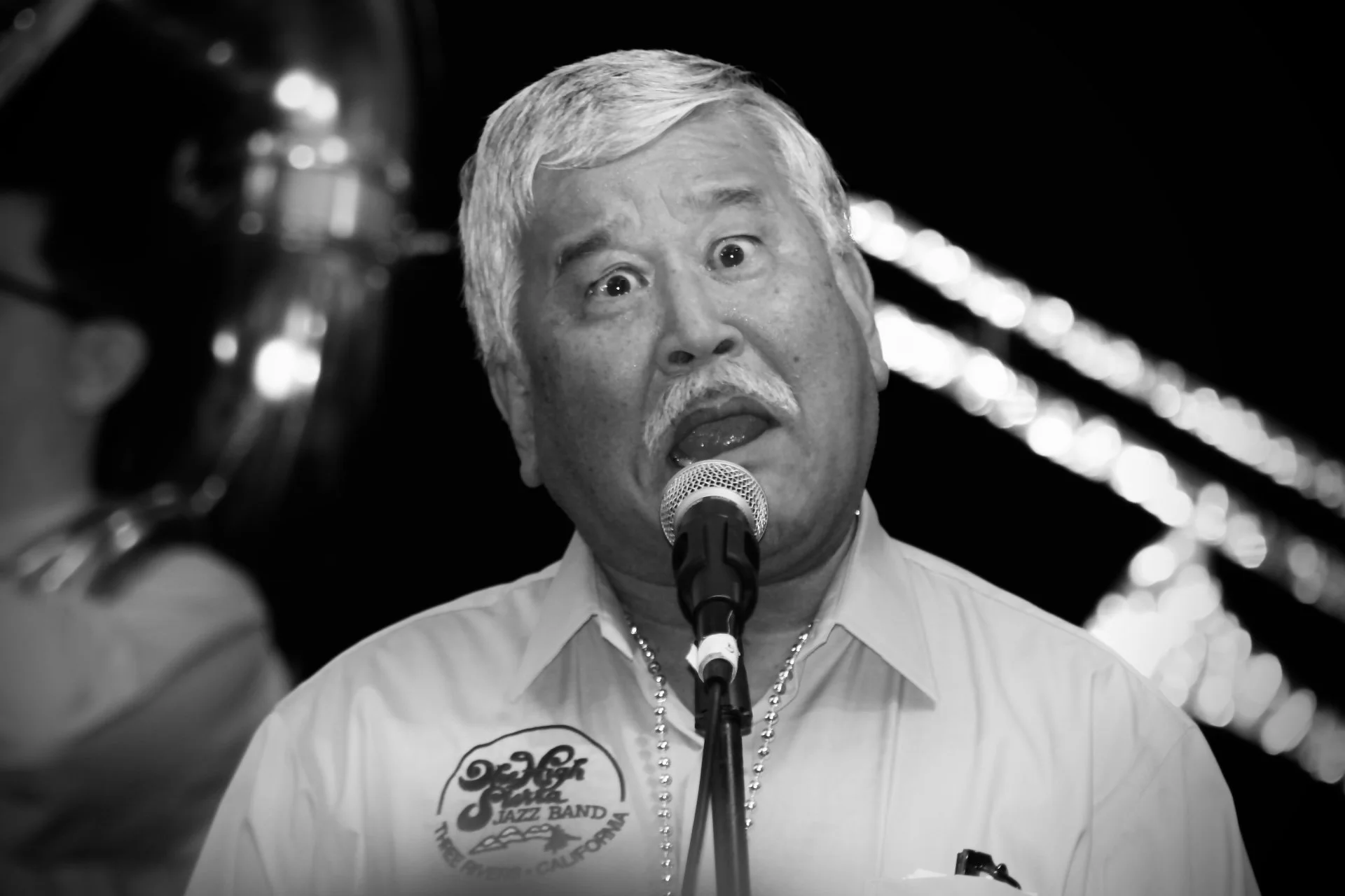 Black and white photo of an older man singing into a microphone, wearing a light-colored shirt with a logo for a jazz band on it, and a necklace, standing in front of a dark background with blurred lights.
