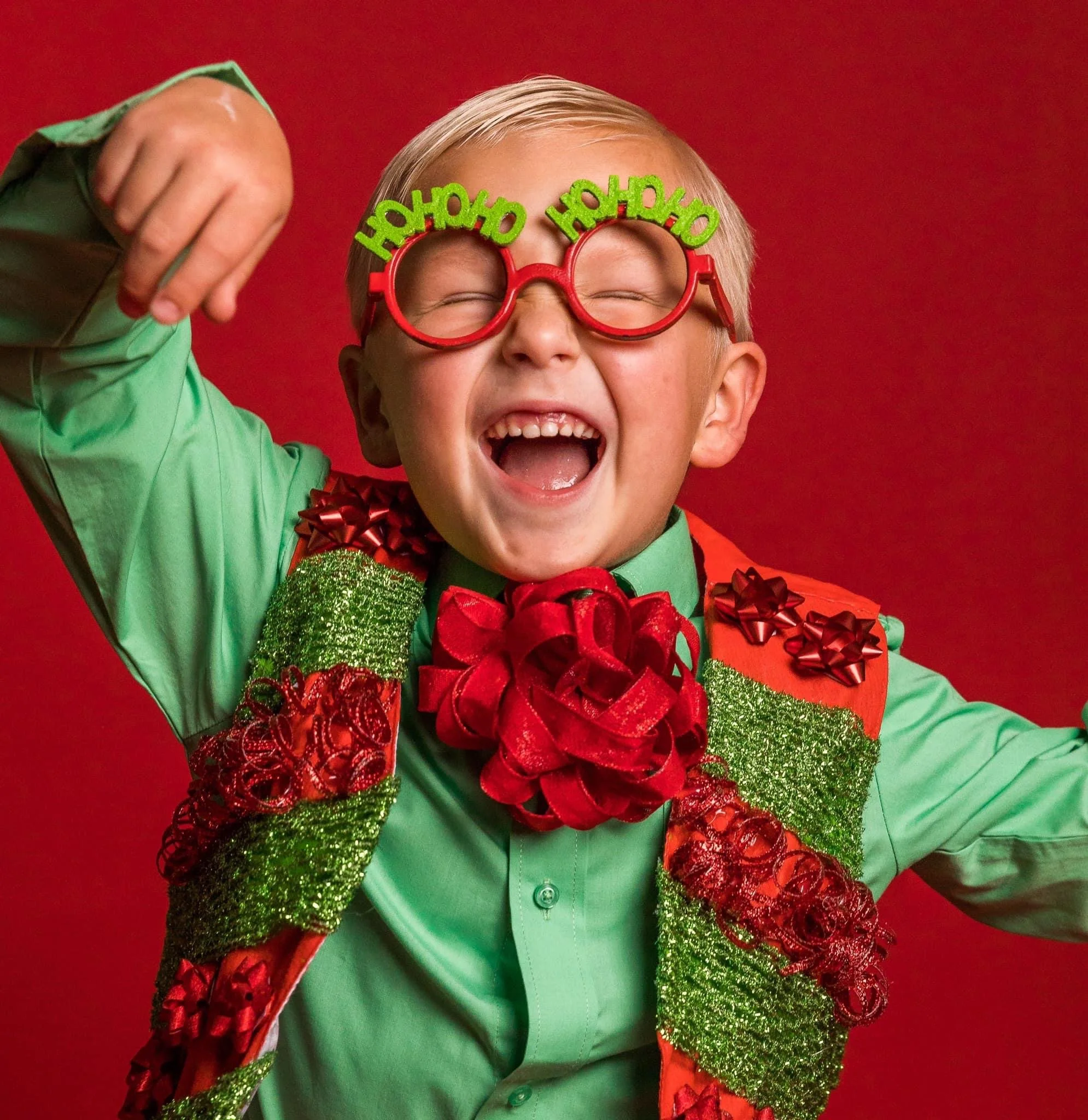 Close-up of a young boy laughing, wearing red glasses with 'Oh What Fun' written on top, surrounded by Christmas decorations including green glittery garland and red bows, red background, and a green shirt.