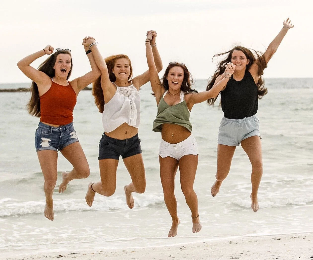 Four young women joyfully jumping on the beach holding hands, with the ocean and sky in the background.
