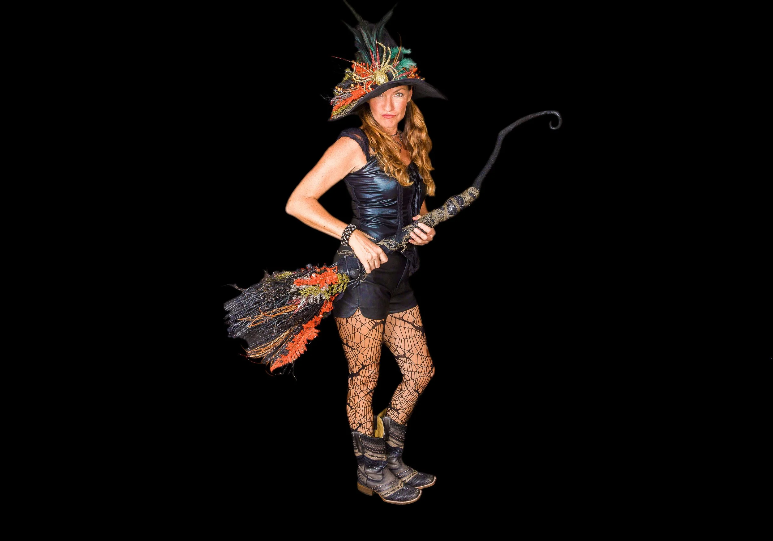 Woman dressed as a witch with a large, decorated hat, holding a broomstick, wearing fishnet stockings and colorful lace-up boots, against a black background.