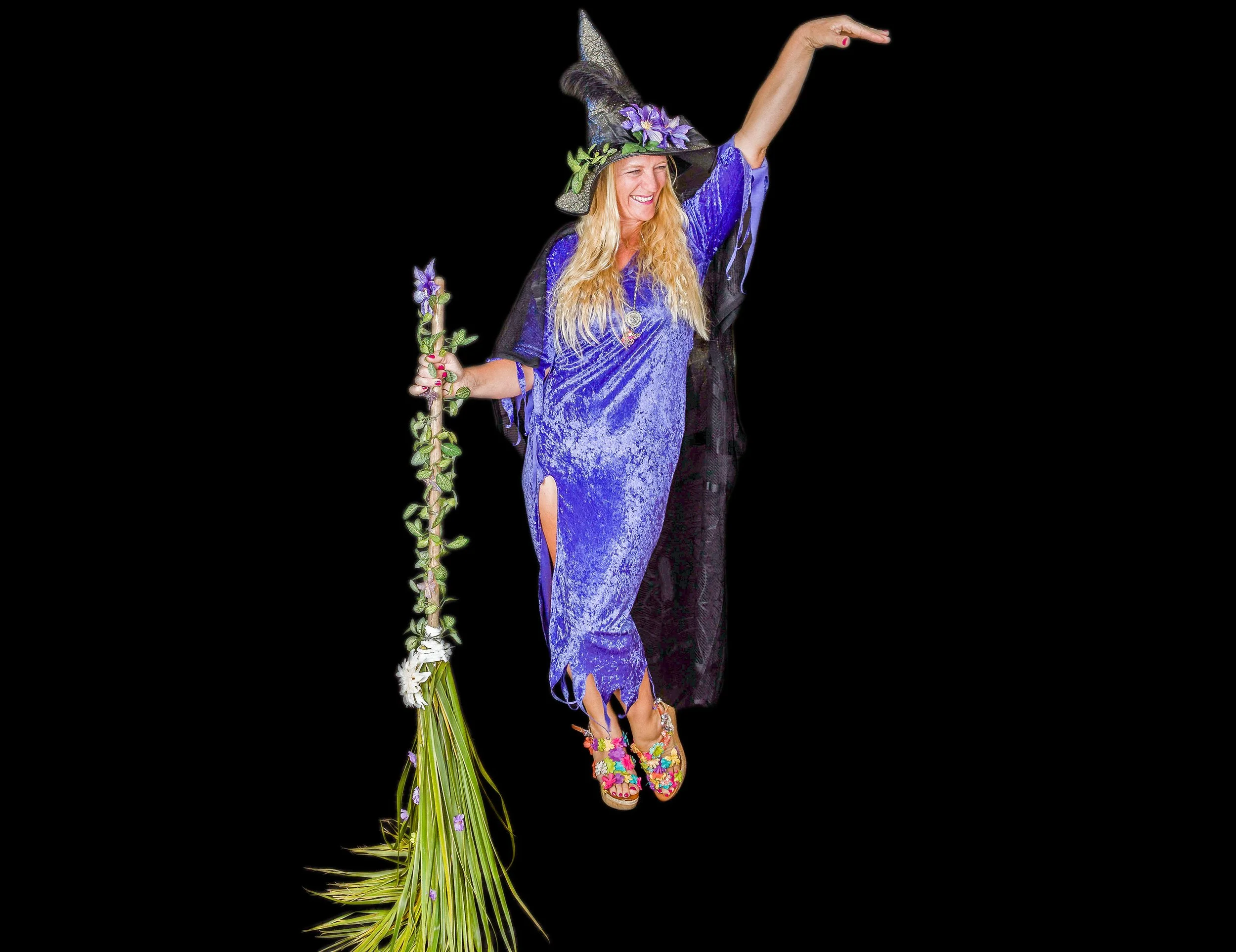 Woman dressed as a witch with a purple dress and witch hat, smiling, holding a purple flower and a bouquet of flowers, standing against a black background.