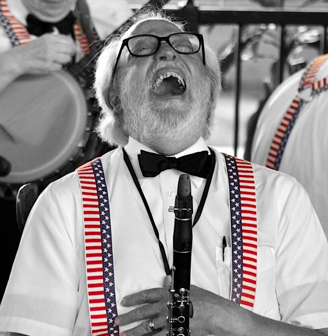 An elderly man with white hair, beard, and glasses, dressed in formal attire with a black bow tie, American flag-themed suspenders, and holding a clarinet while singing passionately.