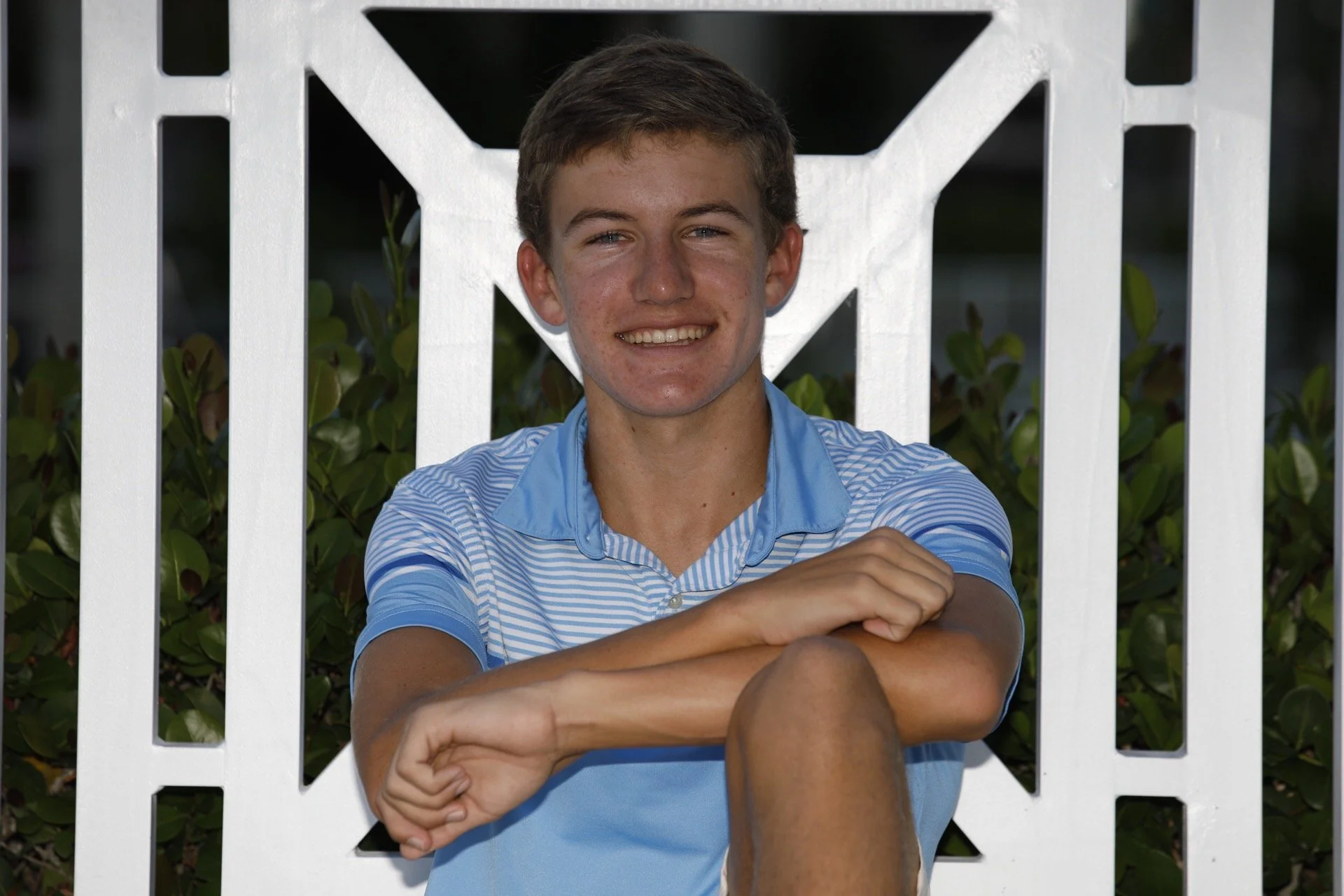 A young person with short brown hair and a striped blue shirt sitting outdoors, smiling at the camera, with their arms resting on their knee, in front of a white decorative fence and green foliage.