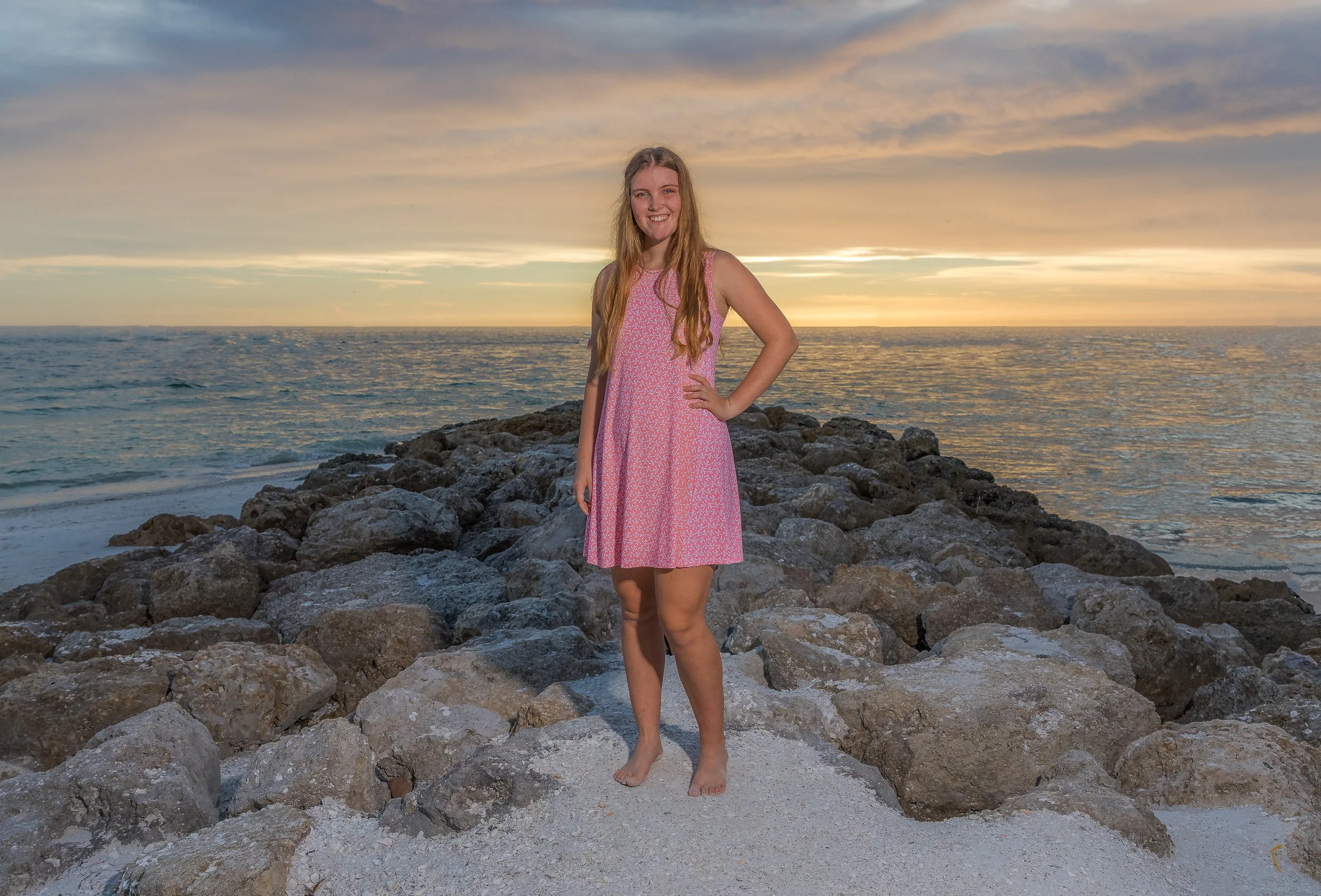 A woman in a pink dress standing on rocks at the beach during sunset.