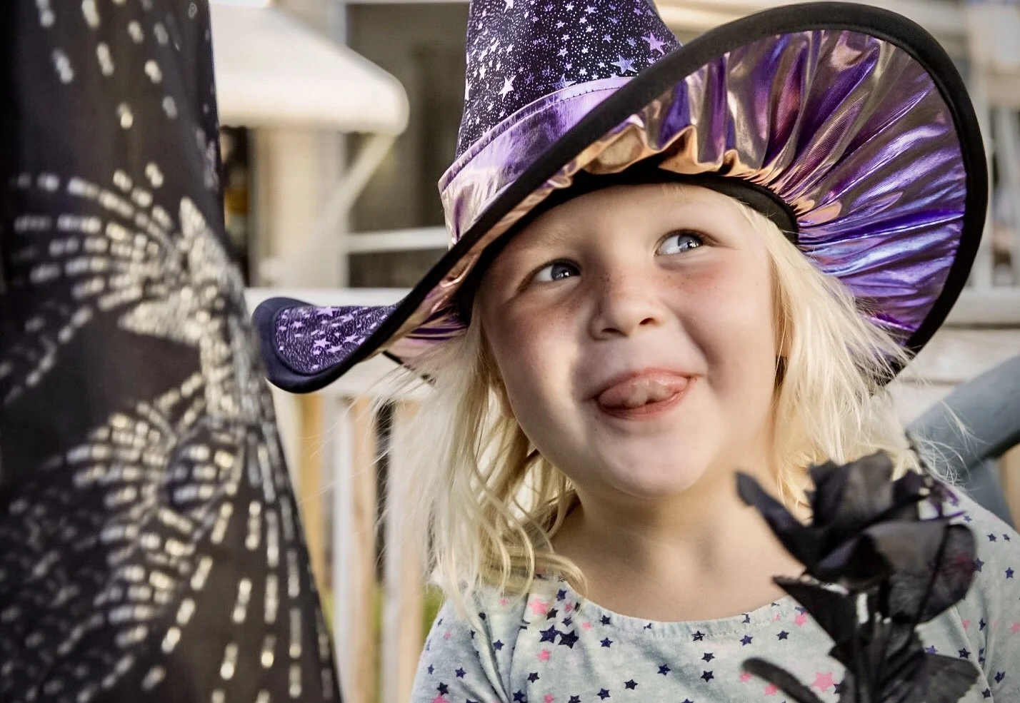 A young girl wearing a purple witch hat with star patterns, sticking out her tongue while holding a black flower, outdoors in daytime.