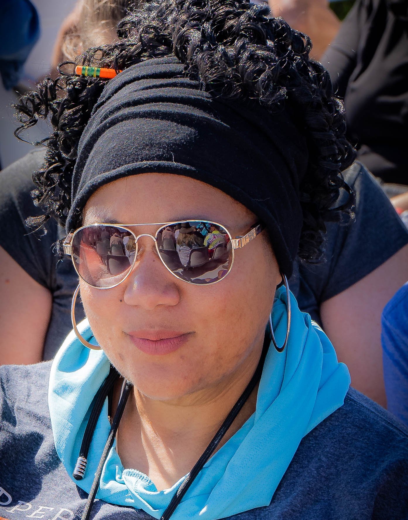 Close-up of a woman wearing sunglasses, a black headscarf, a blue jacket, and earbuds, with a reflection of a group of people in her sunglasses.