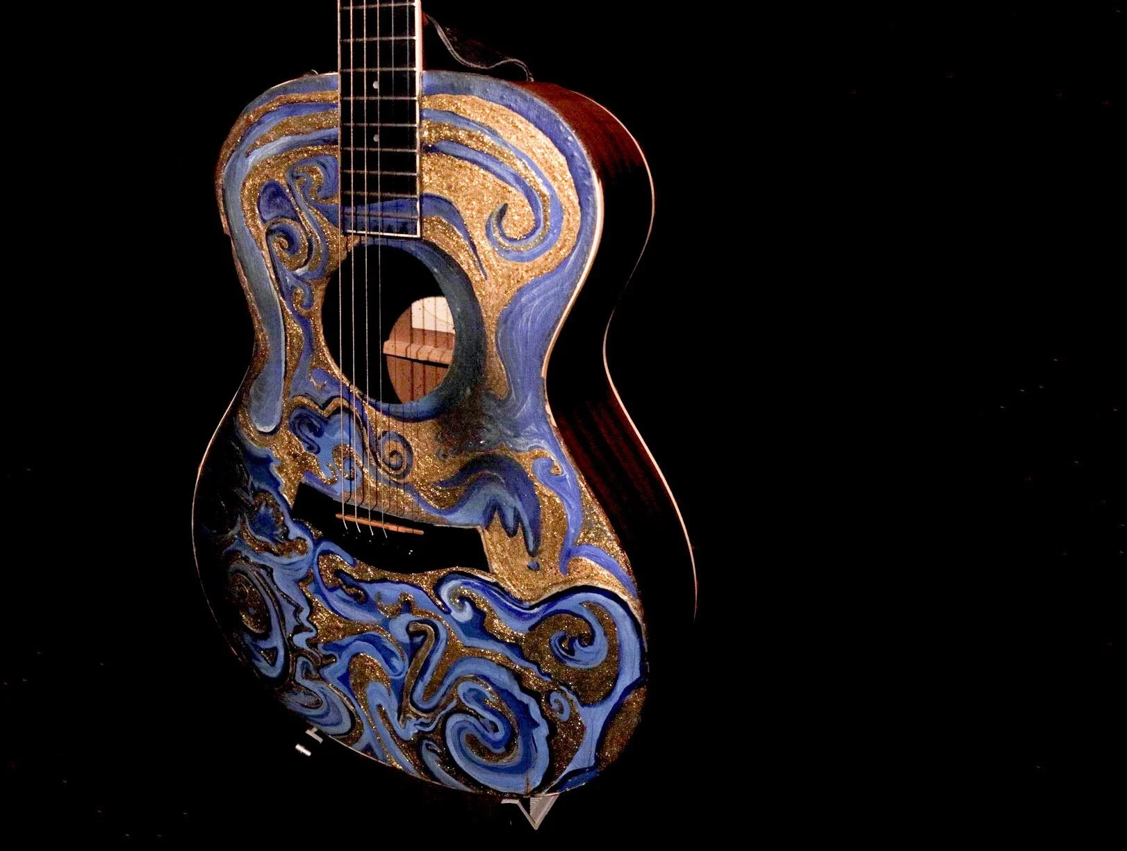 Decorative acoustic guitar with swirling blue and gold patterns on a black background, reflecting on a shiny surface.