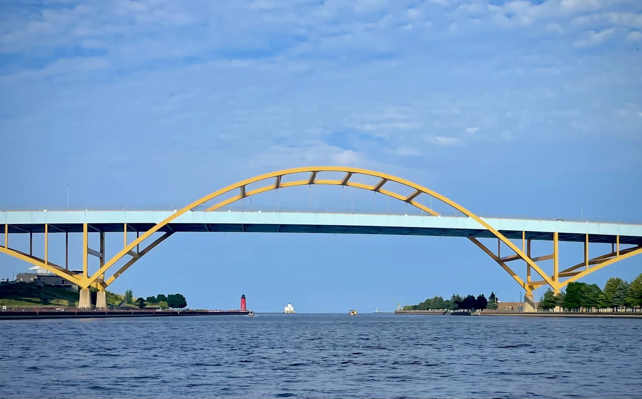 Yellow arched bridge over water with a sky filled with clouds, a lighthouse, and some trees in the background.
