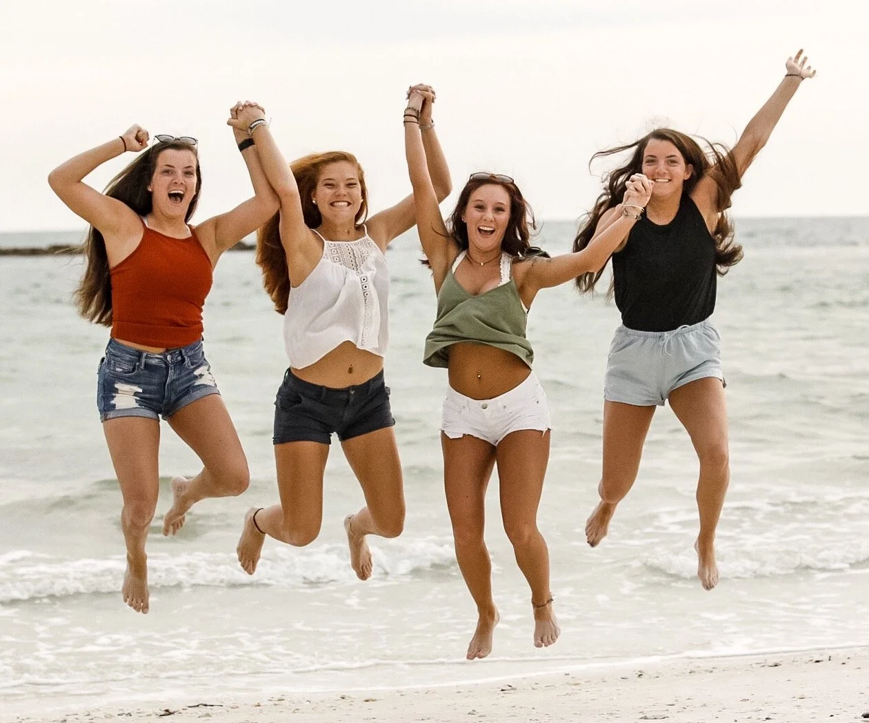 Four young women jumping and holding hands on the beach with ocean in the background.