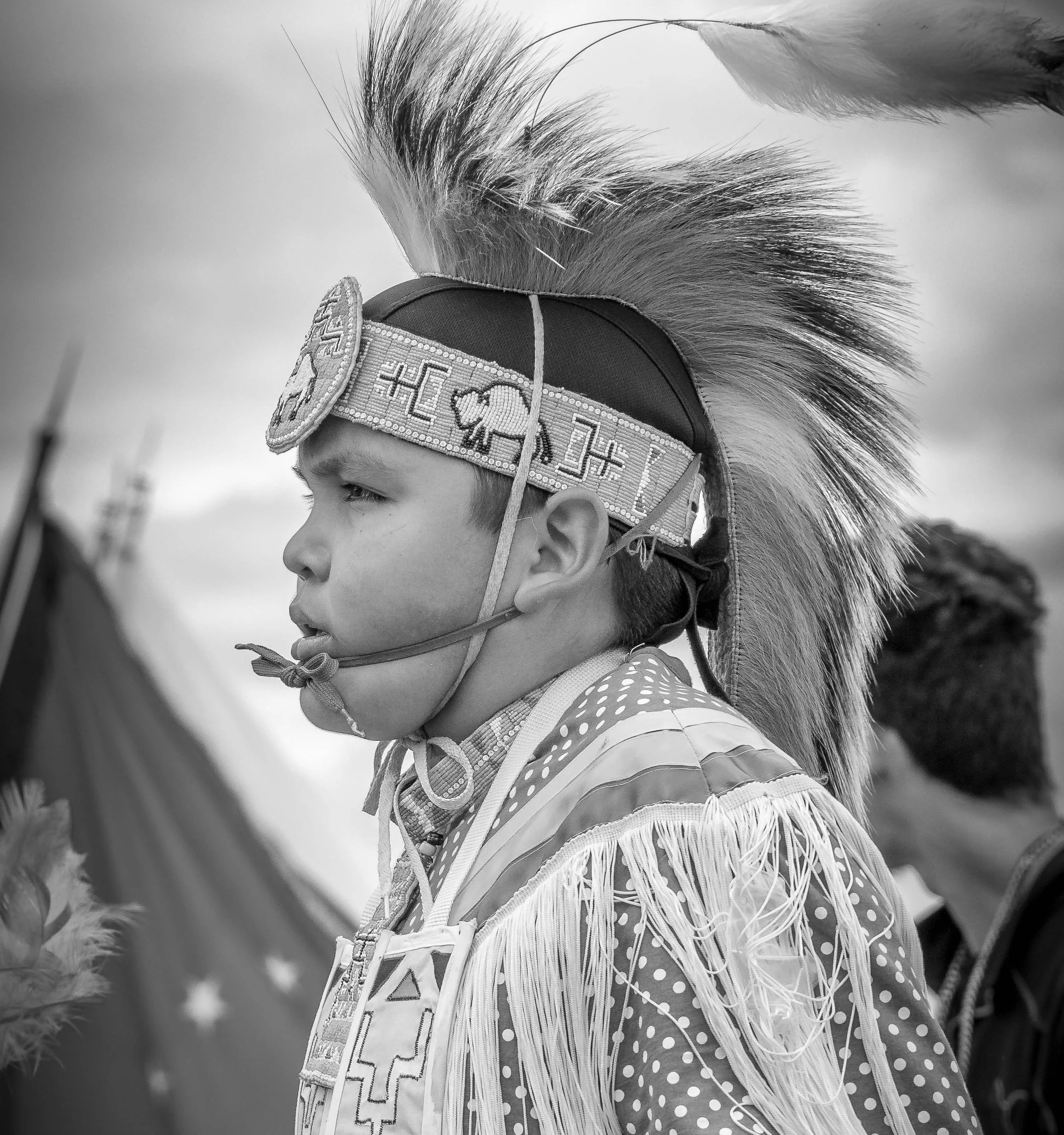 A young person dressed in traditional Native American attire, including a detailed beaded headband and decorated clothing with fringe and geometric pattern, wearing a feathered headdress, during a cultural event or celebration.